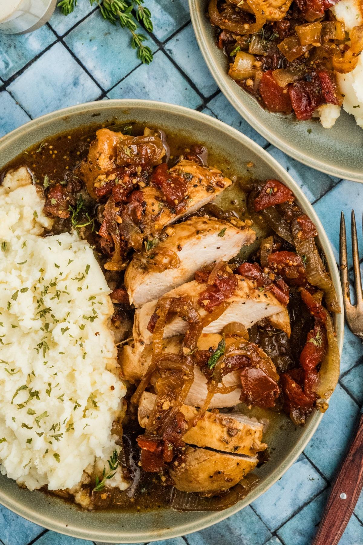 A plate of sliced chicken with stewed tomatoes and onions, served with mashed potatoes and herbs.