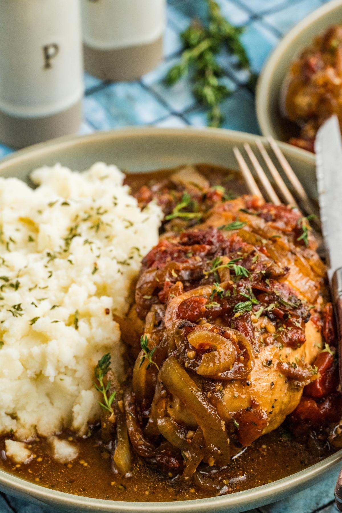 A plate of mashed potatoes and braised chicken with onions, tomatoes, and herbs, served with a fork and knife.