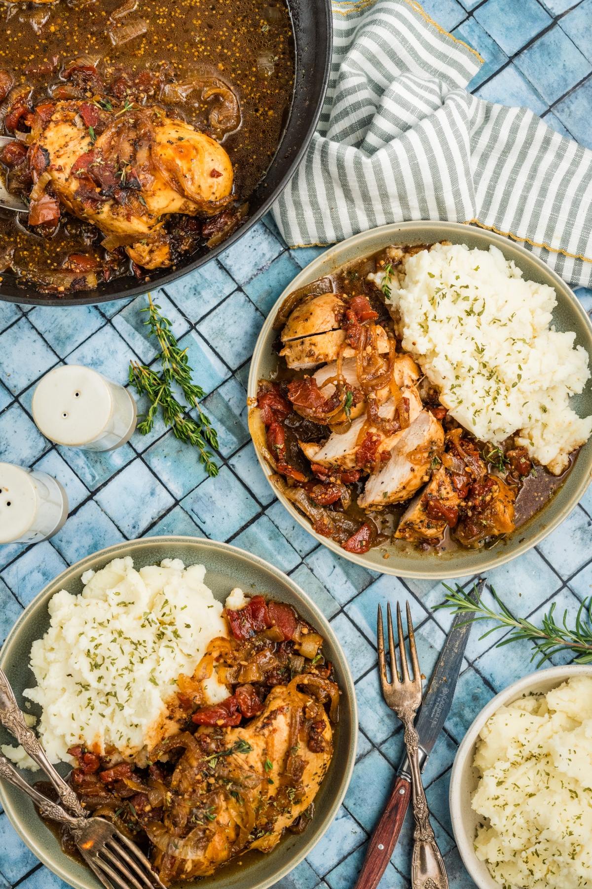 Two plates of chicken with sauce and mashed potatoes, a skillet of chicken, napkin, herbs, forks, and salt shakers.