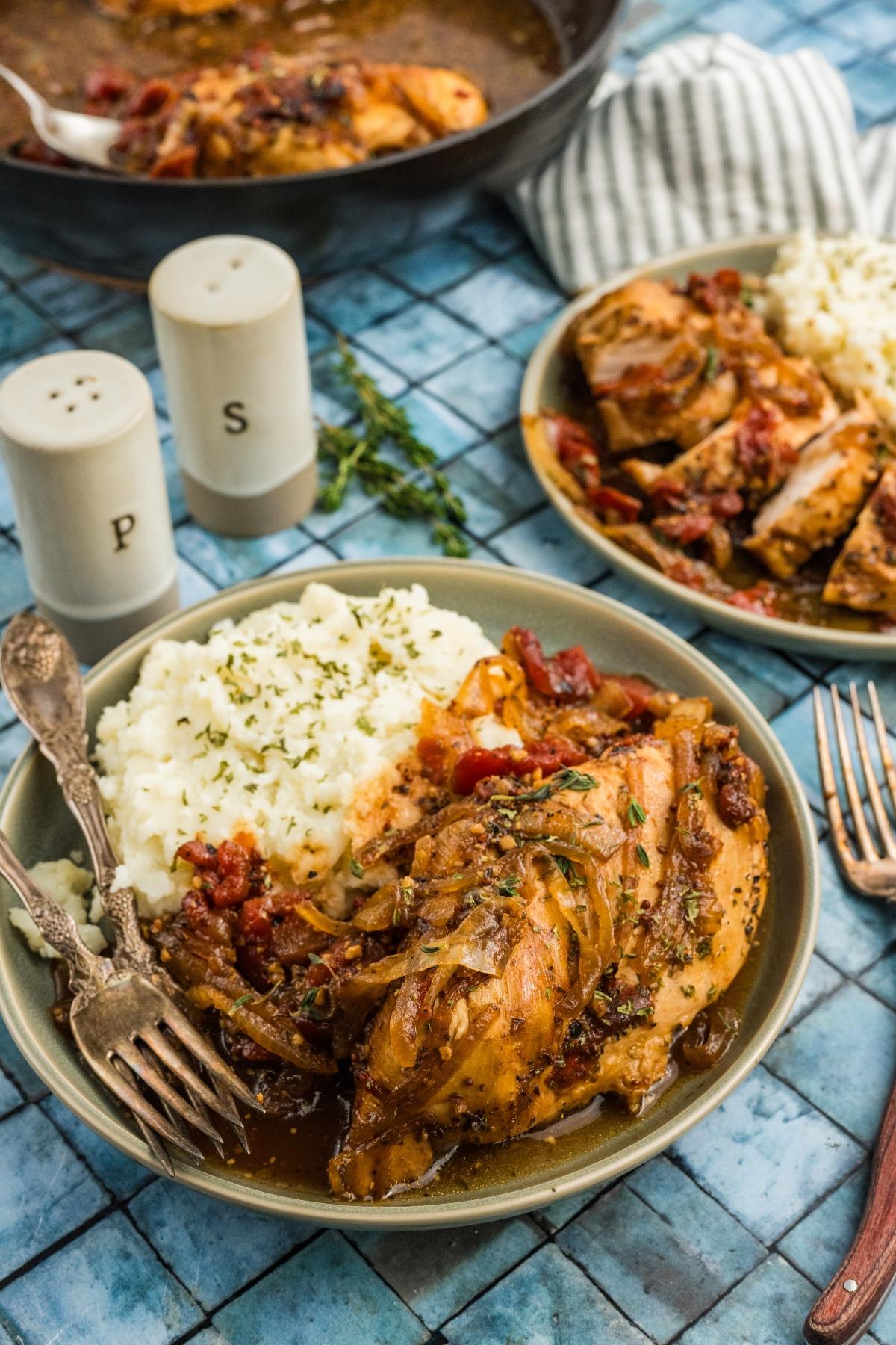 A plate of chicken with onions, mashed potatoes, and herbs, set on a blue tile table with cutlery and seasonings.