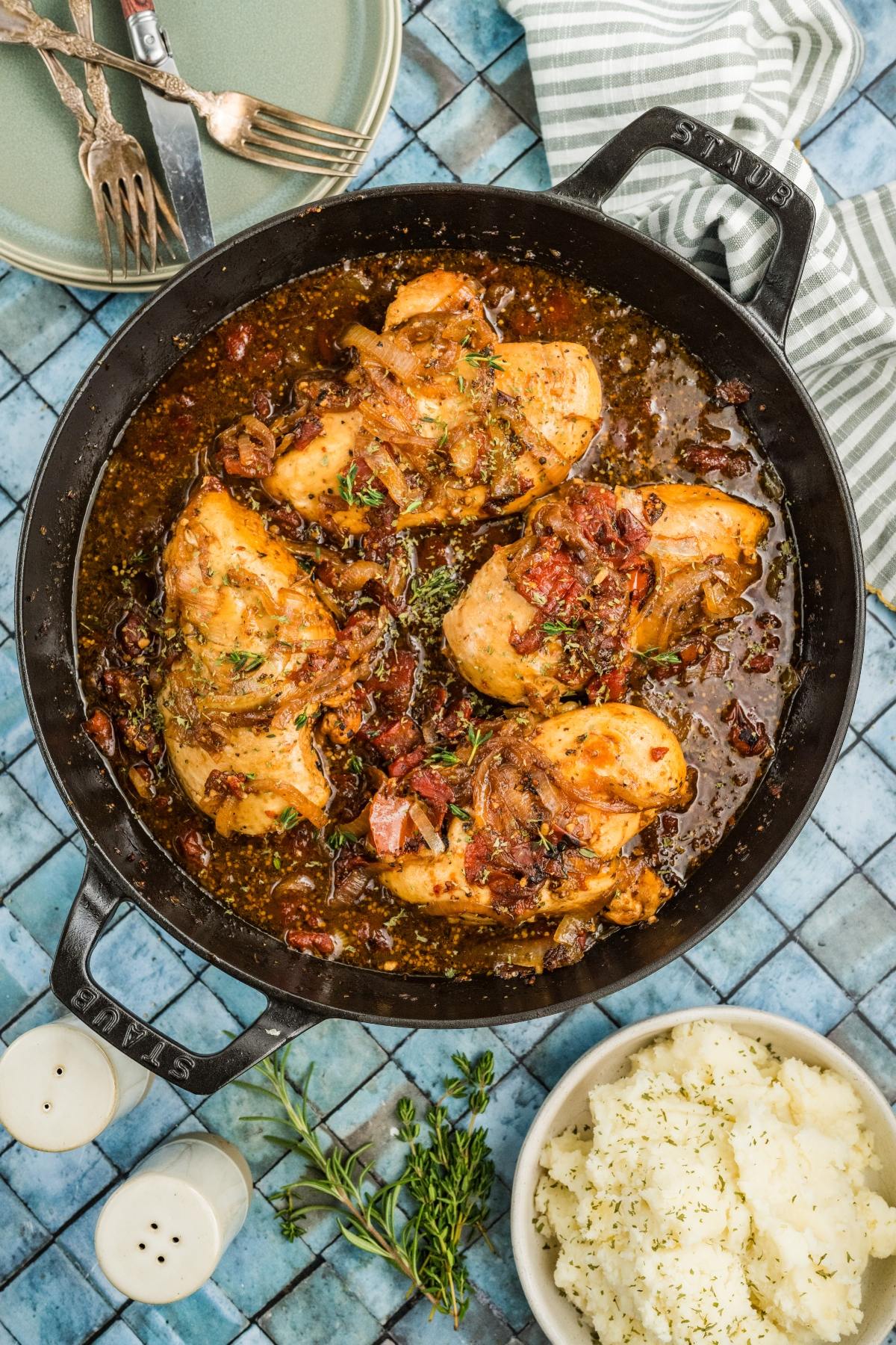 A skillet of braised chicken with herbs, served with mashed potatoes on a blue tiled surface.