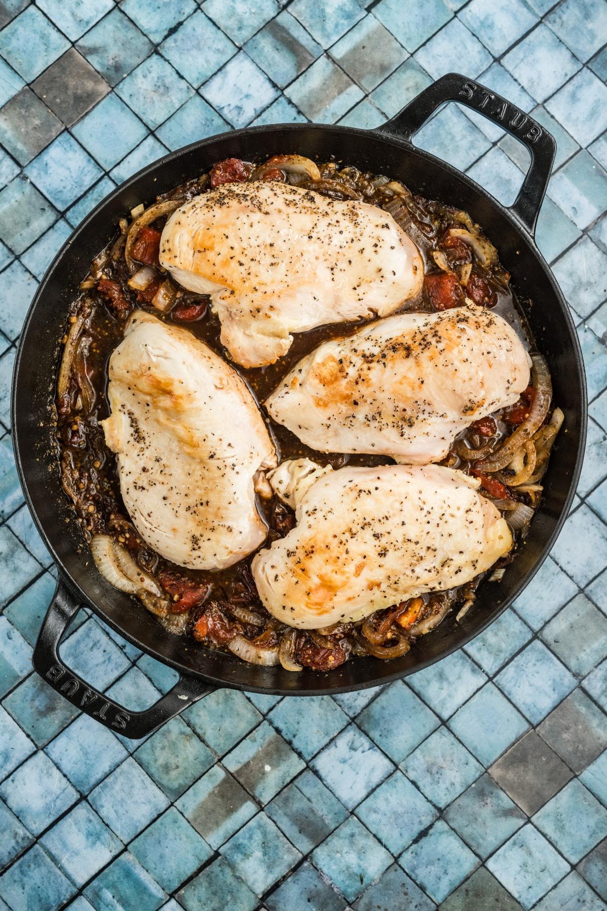Four seasoned chicken breasts cooking in a skillet with onions and tomatoes on a blue tiled surface.
