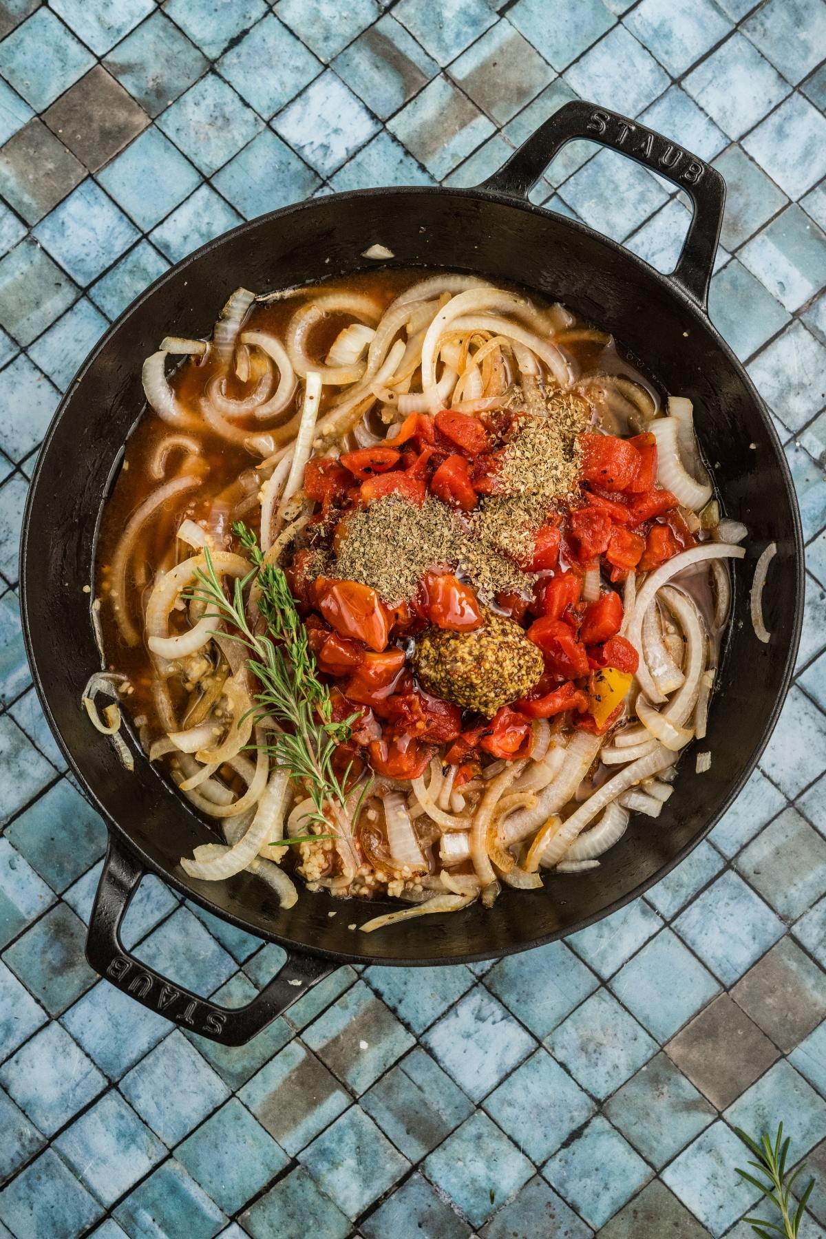 A black pan with onions, chopped tomatoes, herbs, and spices on a blue-tiled surface.
