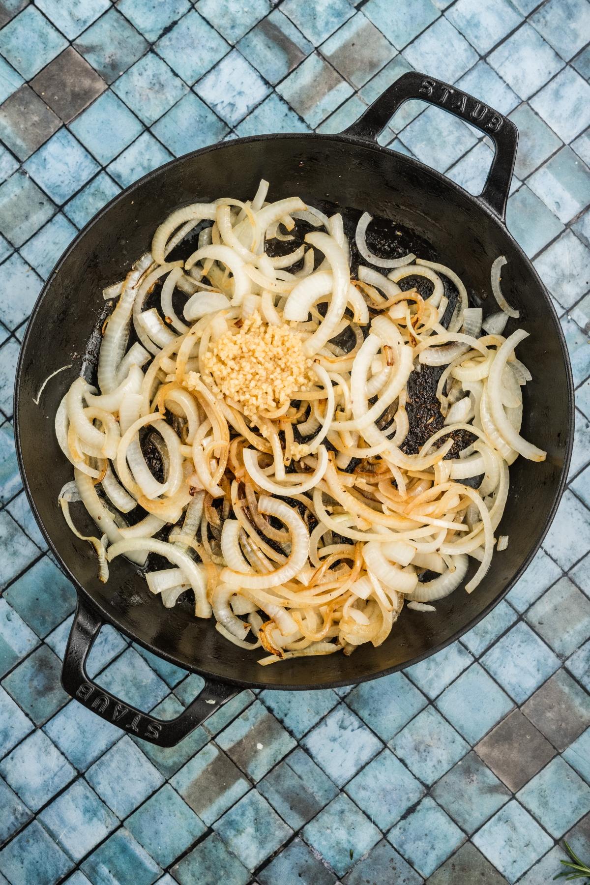 Sliced onions and minced garlic cooking in a black skillet on a blue tiled surface.