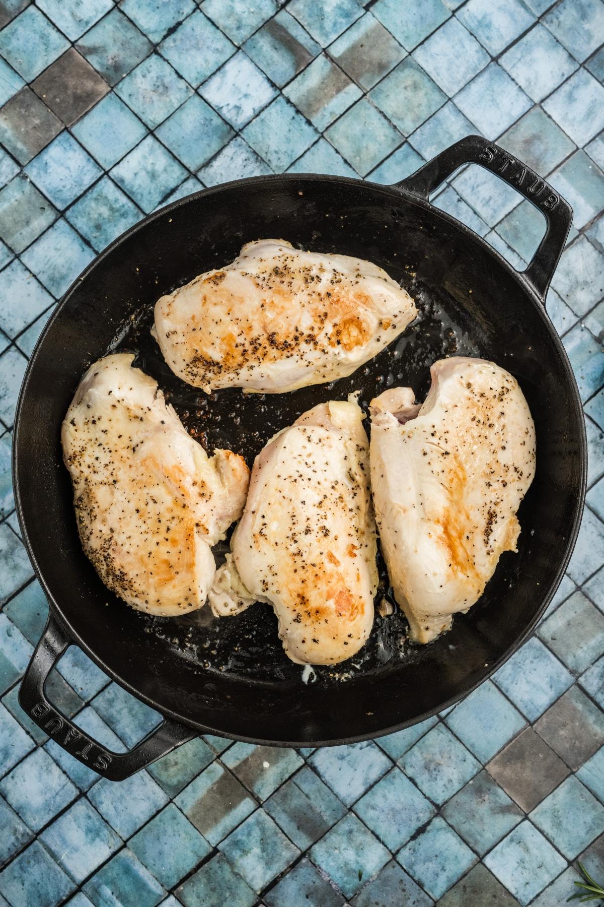 Four seasoned chicken breasts cooking in a black skillet on a blue tiled surface.