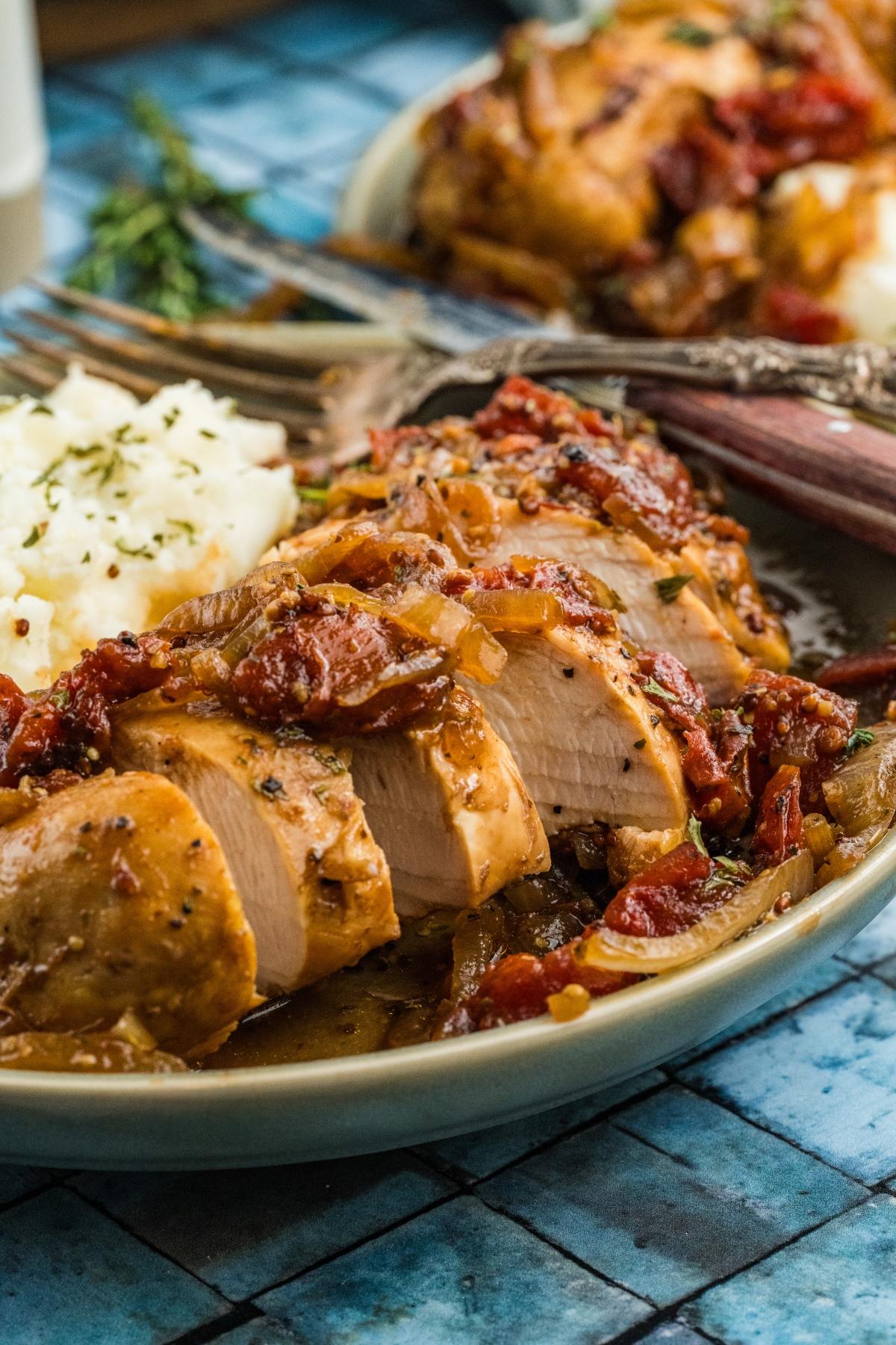 Sliced chicken breast with tomatoes and onions, served with mashed potatoes on a blue-patterned plate.