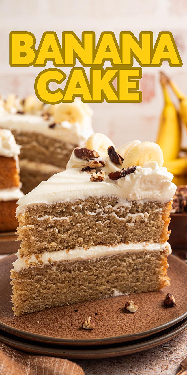 A slice of banana cake with creamy frosting and fresh banana slices on top, surrounded by ripe bananas in the background.