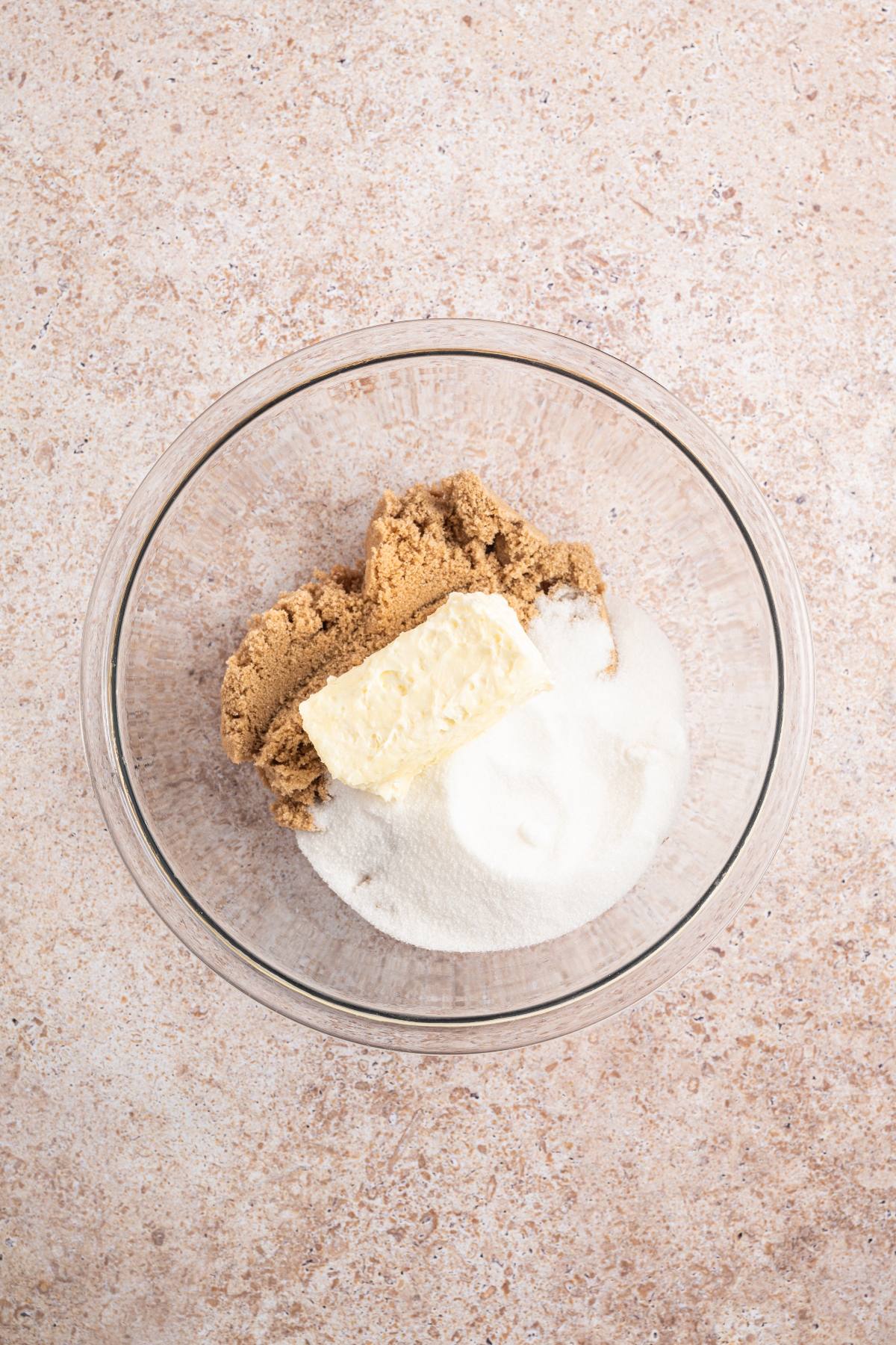 A glass bowl with butter, brown sugar, and white sugar on a beige countertop.