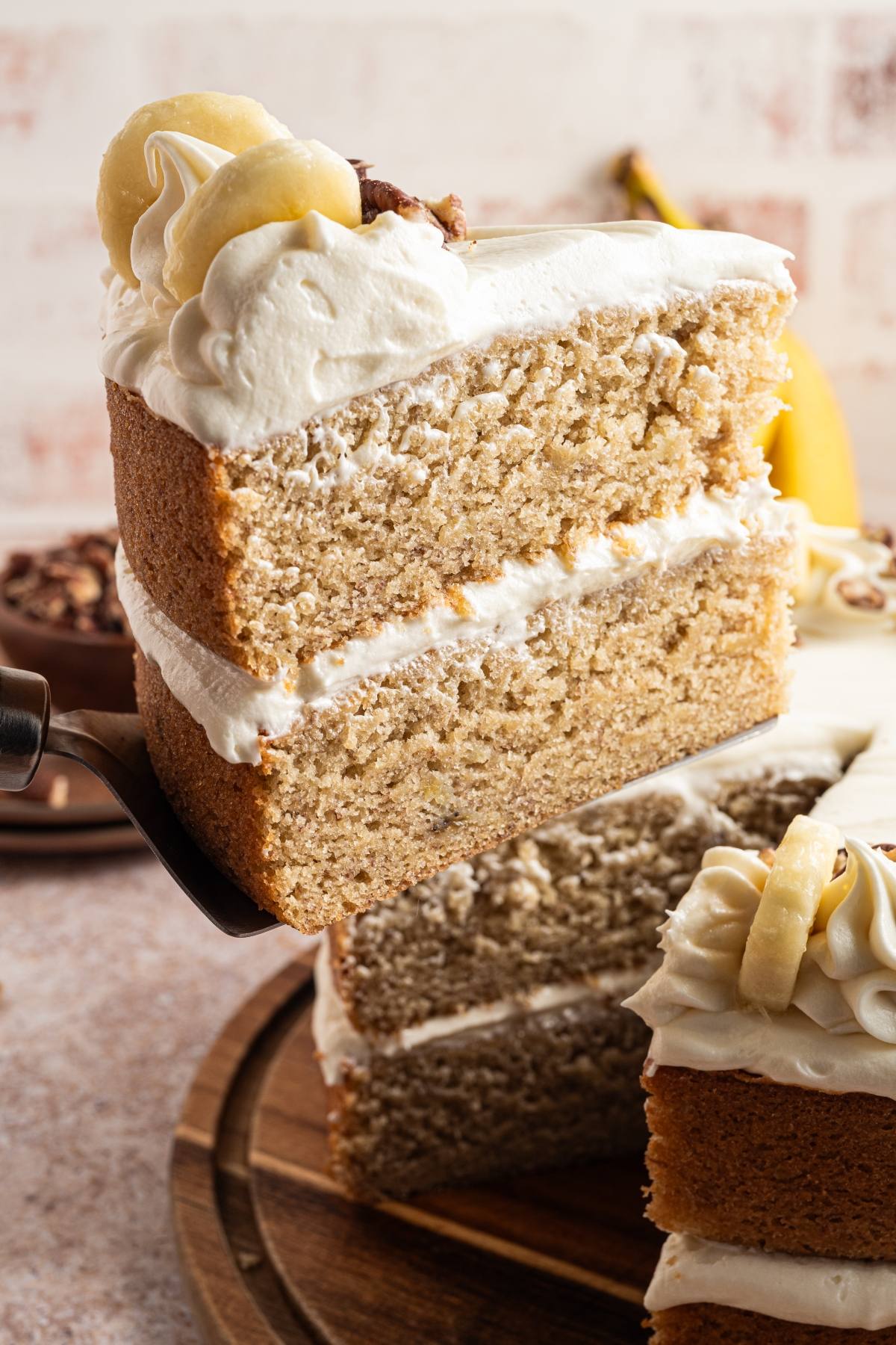 A slice of banana cake with white frosting is being lifted from a whole cake, topped with banana slices.