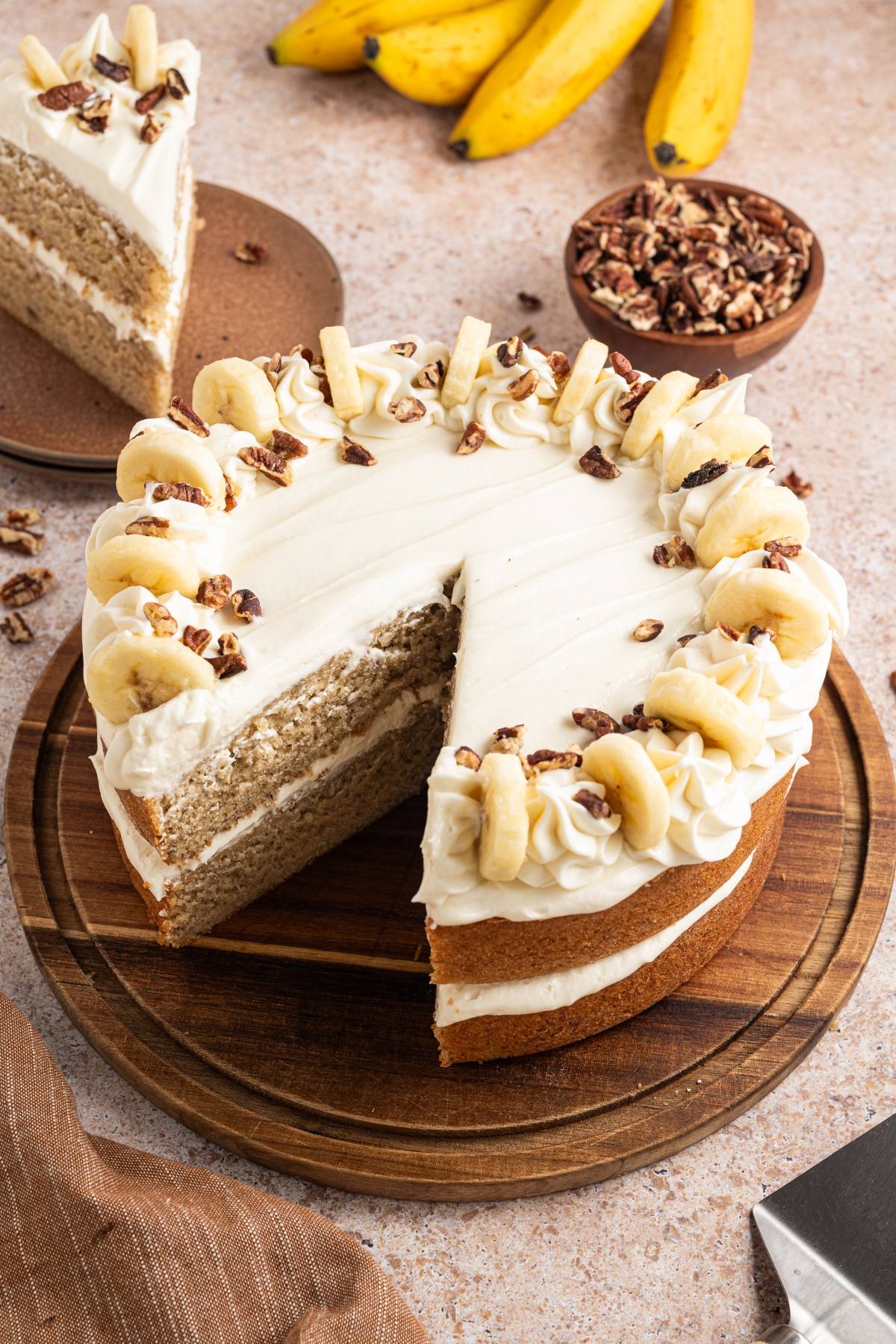 Round banana cake with white frosting, topped with banana slices and nuts, on a wooden board; one slice cut.