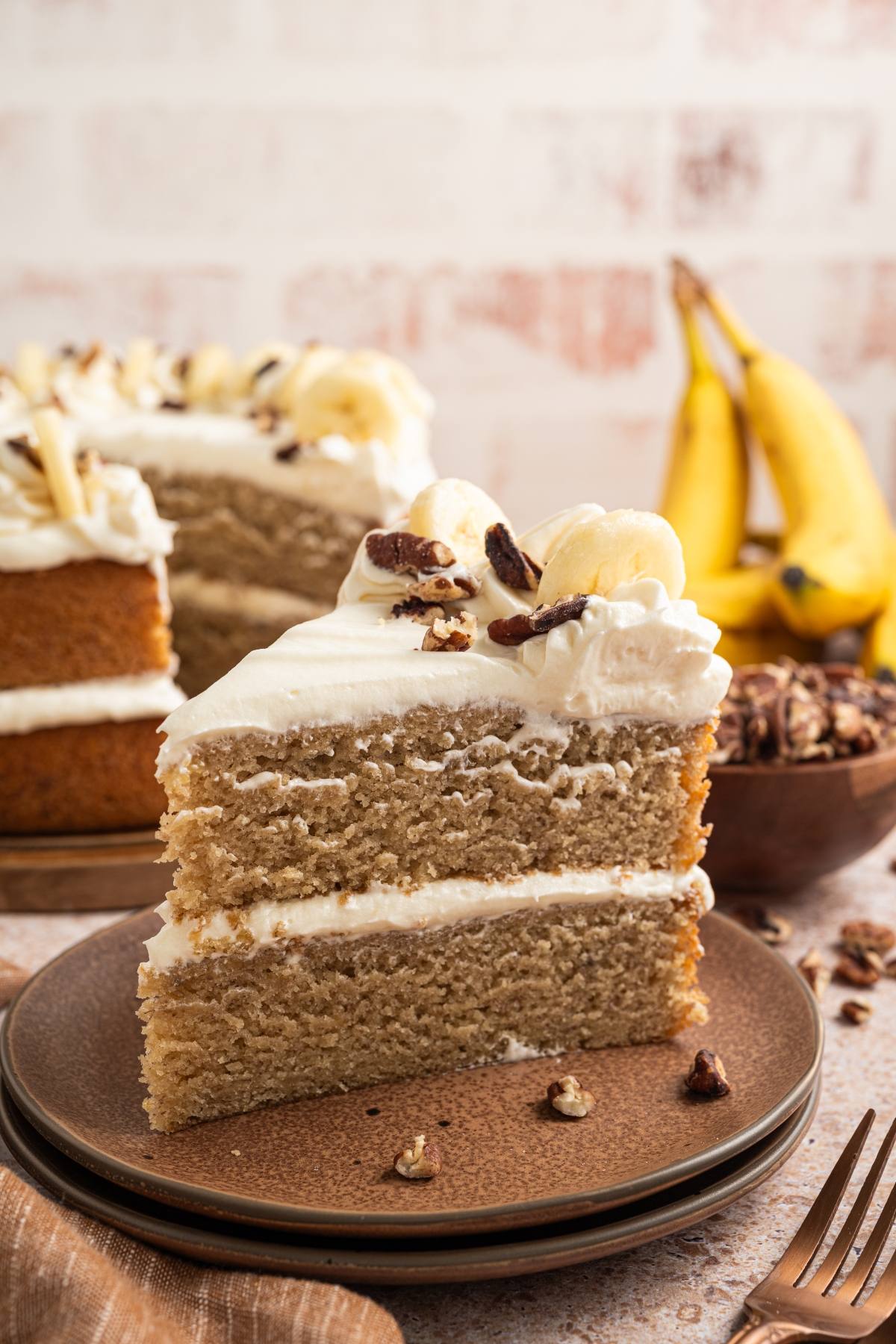 A slice of banana cake with white frosting and nuts on a plate, with bananas and more cake in the background.