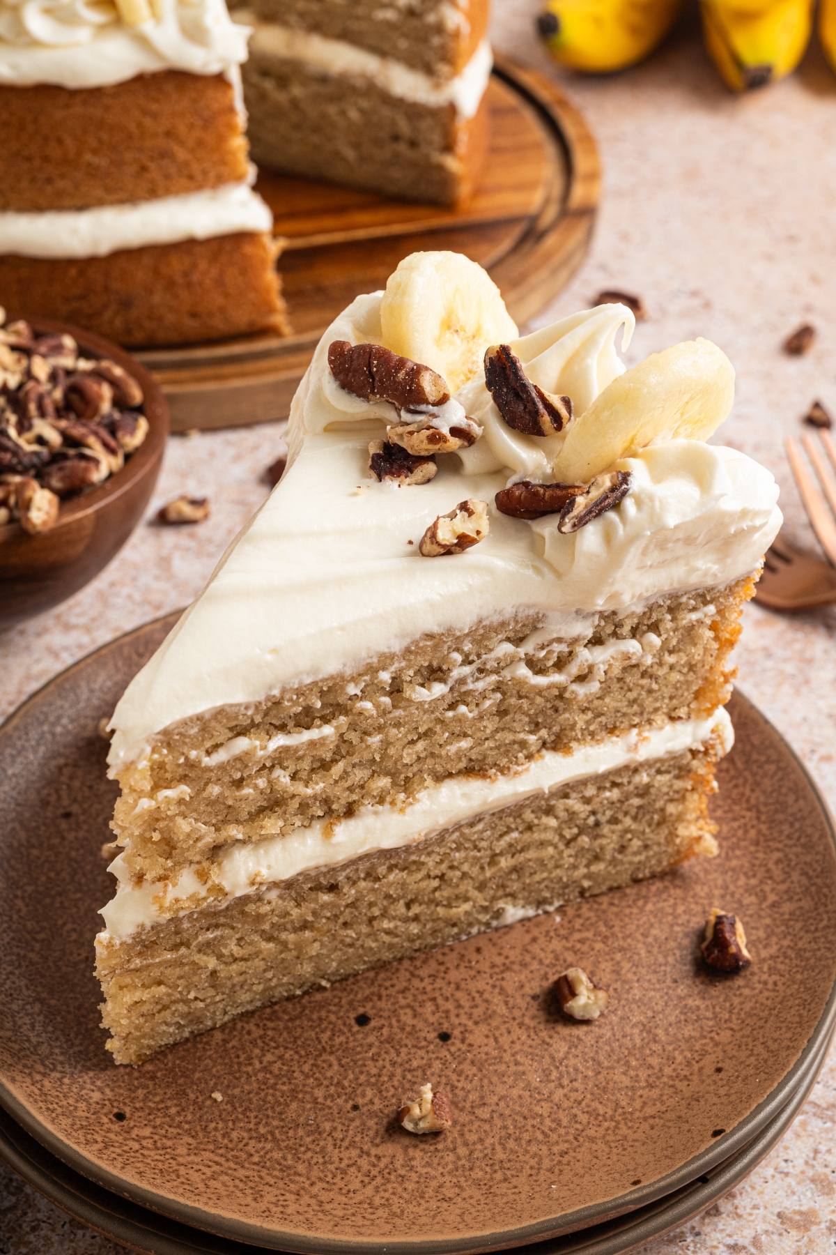 A slice of banana cake with cream frosting, topped with banana slices and nuts, on a brown plate.