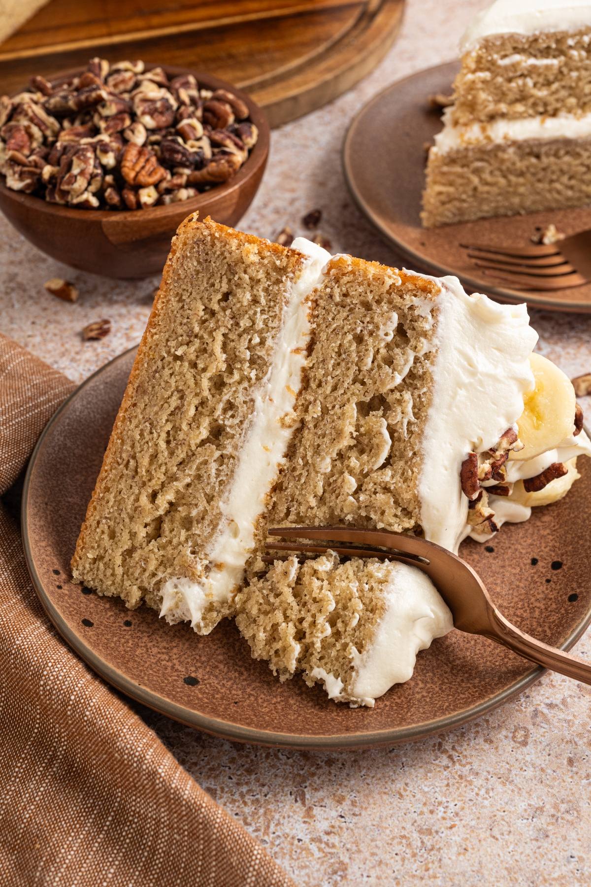 A slice of layered banana cake with cream cheese frosting on a plate, topped with nuts, and a fork beside it.