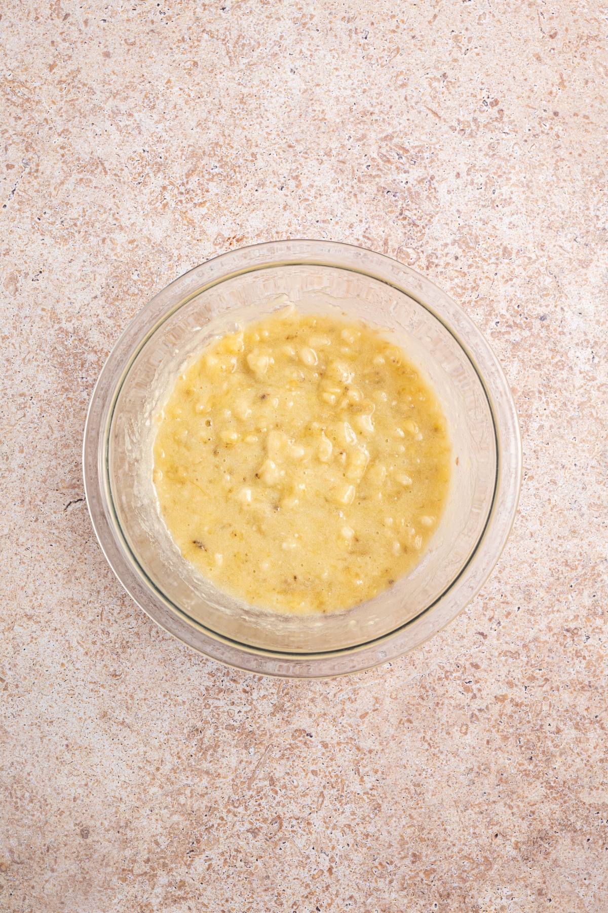 A glass bowl with mashed bananas on a light-colored, textured surface.
