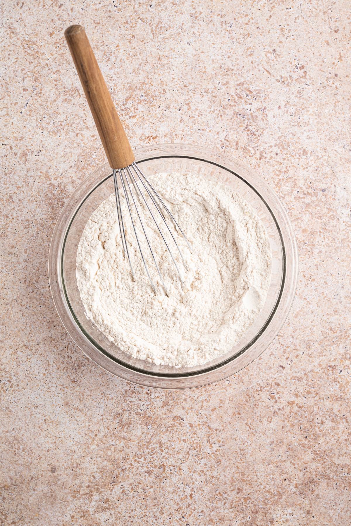 Glass bowl of flour with a metal whisk on a light brown textured countertop.