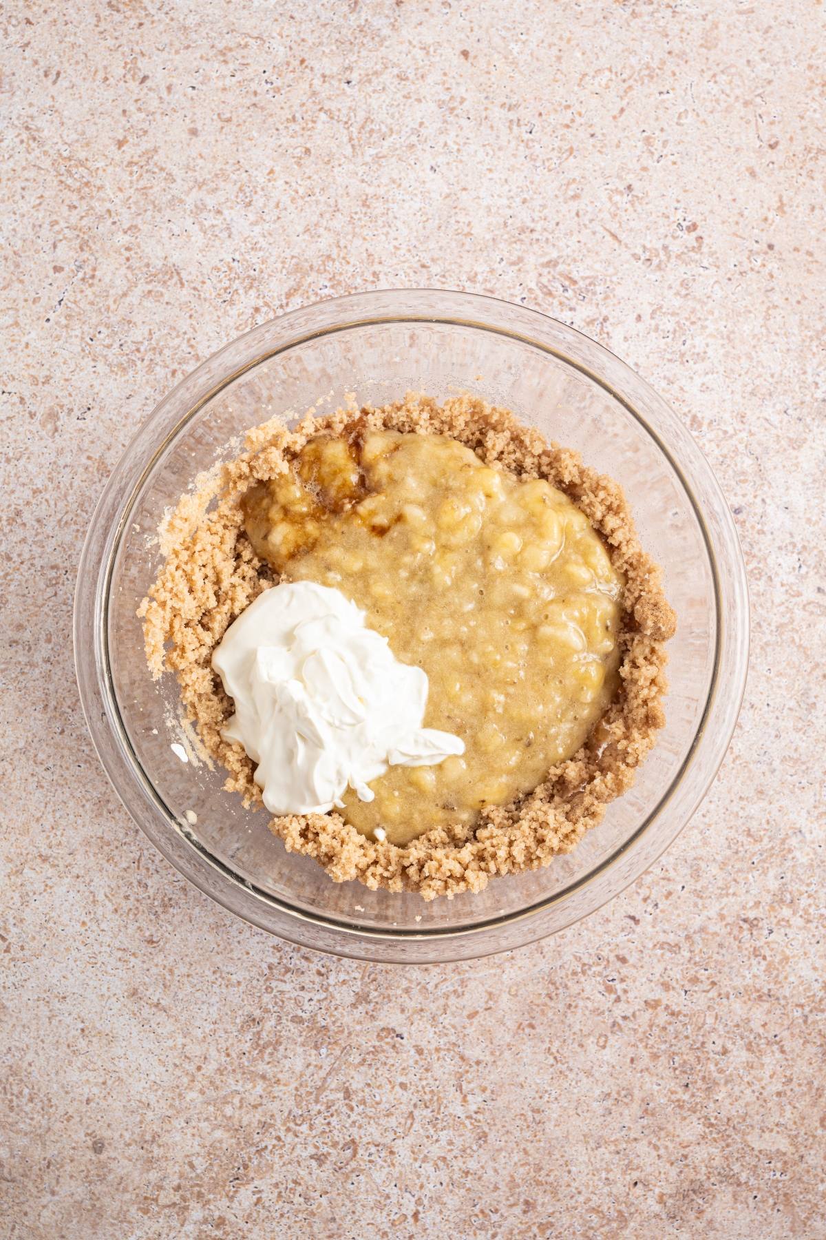 A glass bowl with mashed banana, sour cream, and sugar mixture on a light countertop.