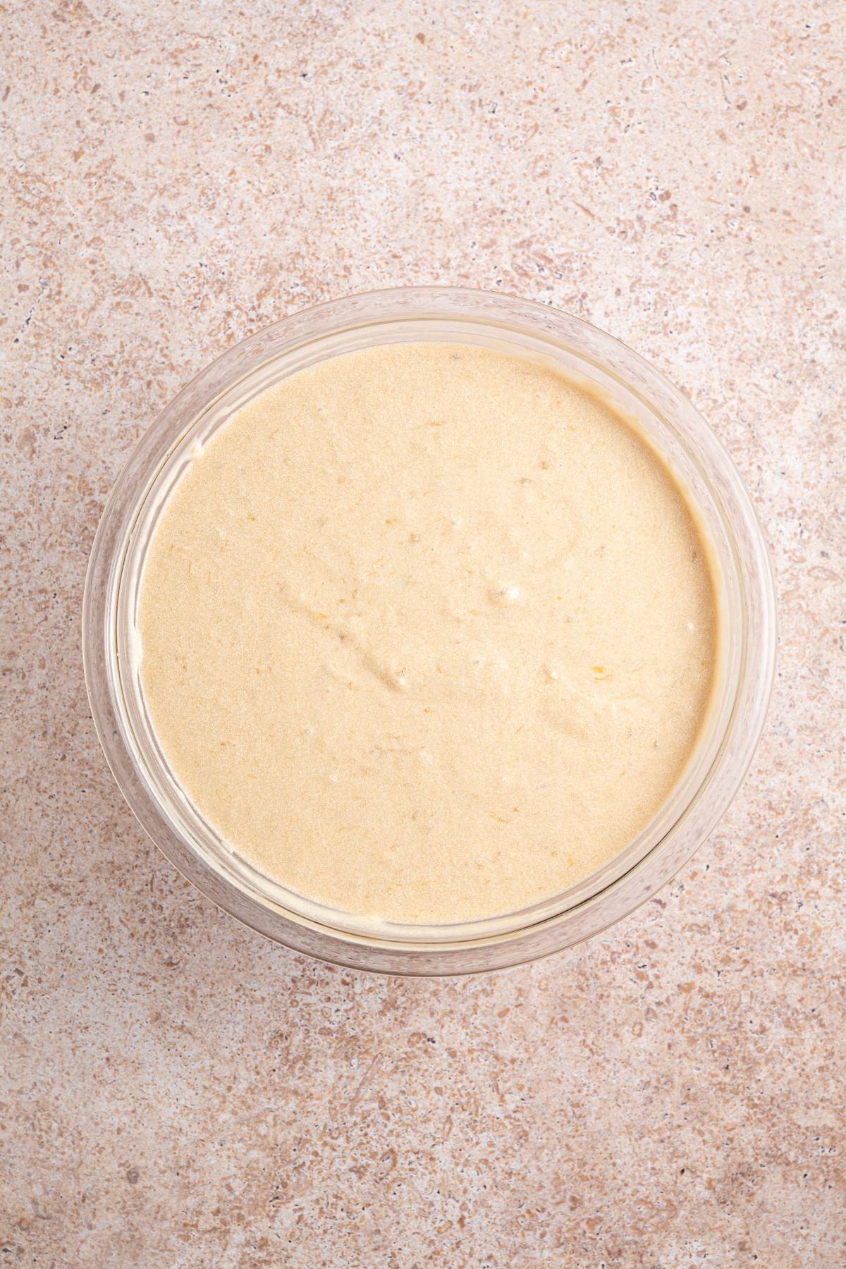 A glass bowl filled with smooth, light beige batter on a speckled countertop.