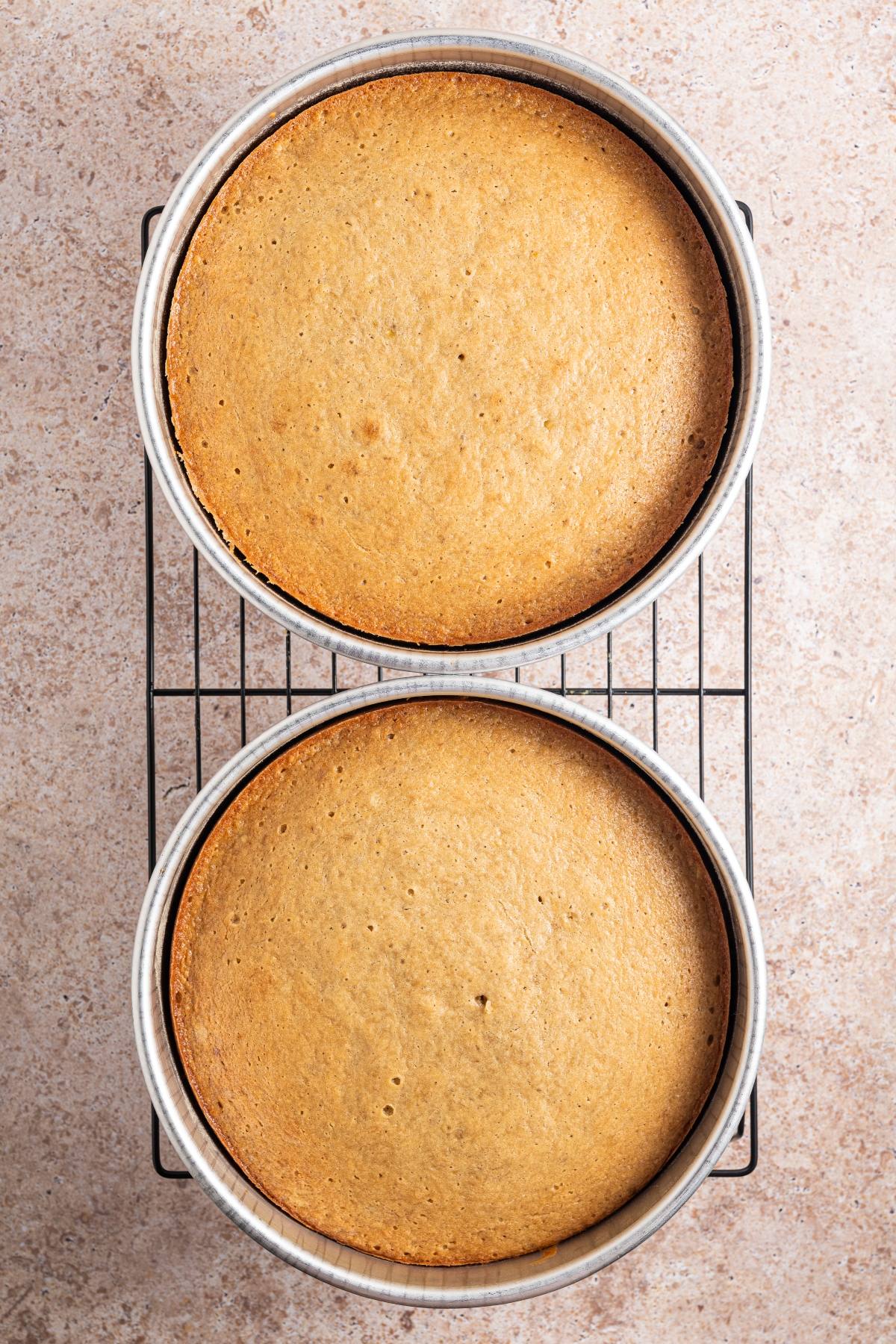 Two round baked cakes in pans cooling on a wire rack over a light-colored countertop.