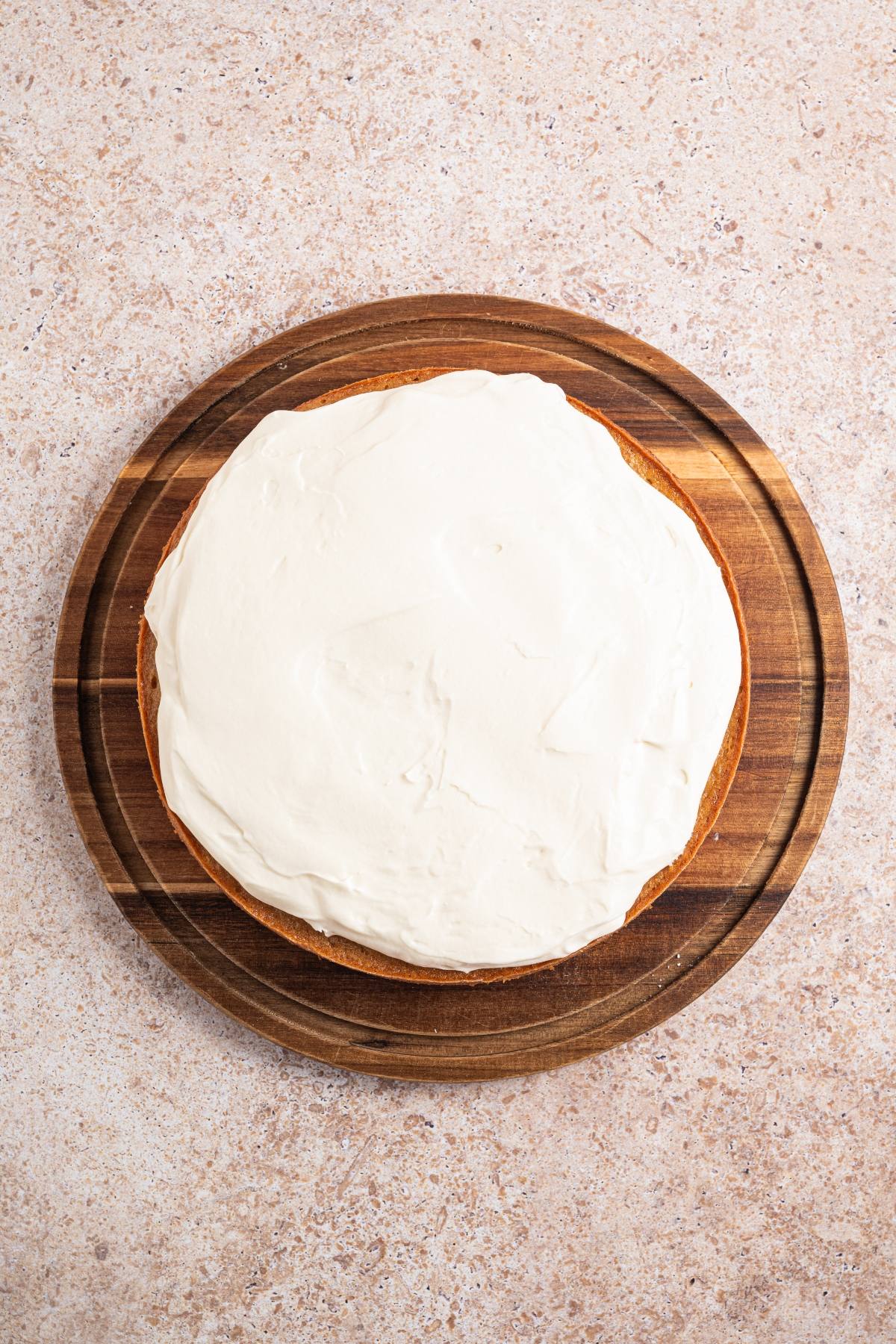 Round cake with white frosting on a wooden board, viewed from above on a light countertop.
