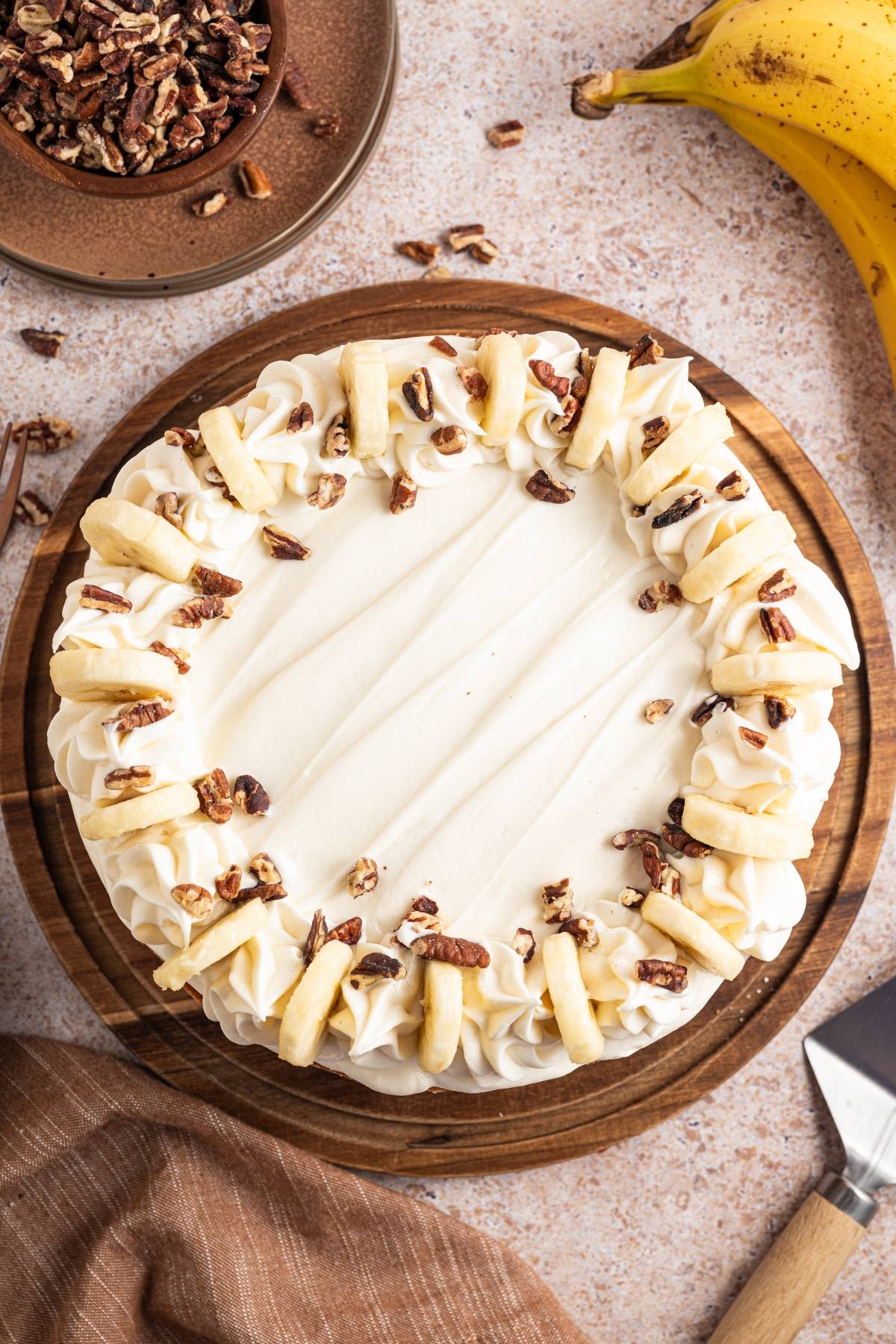 A frosted cake topped with whipped cream, banana slices, and chopped pecans on a wooden serving board.
