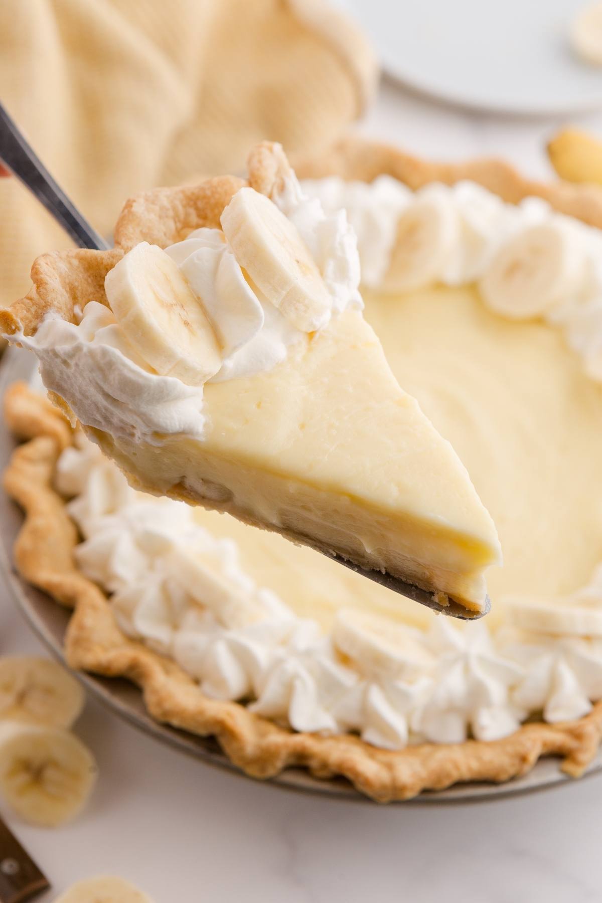 A slice of banana cream pie with whipped cream and fresh banana slices on top is being lifted from the pie.