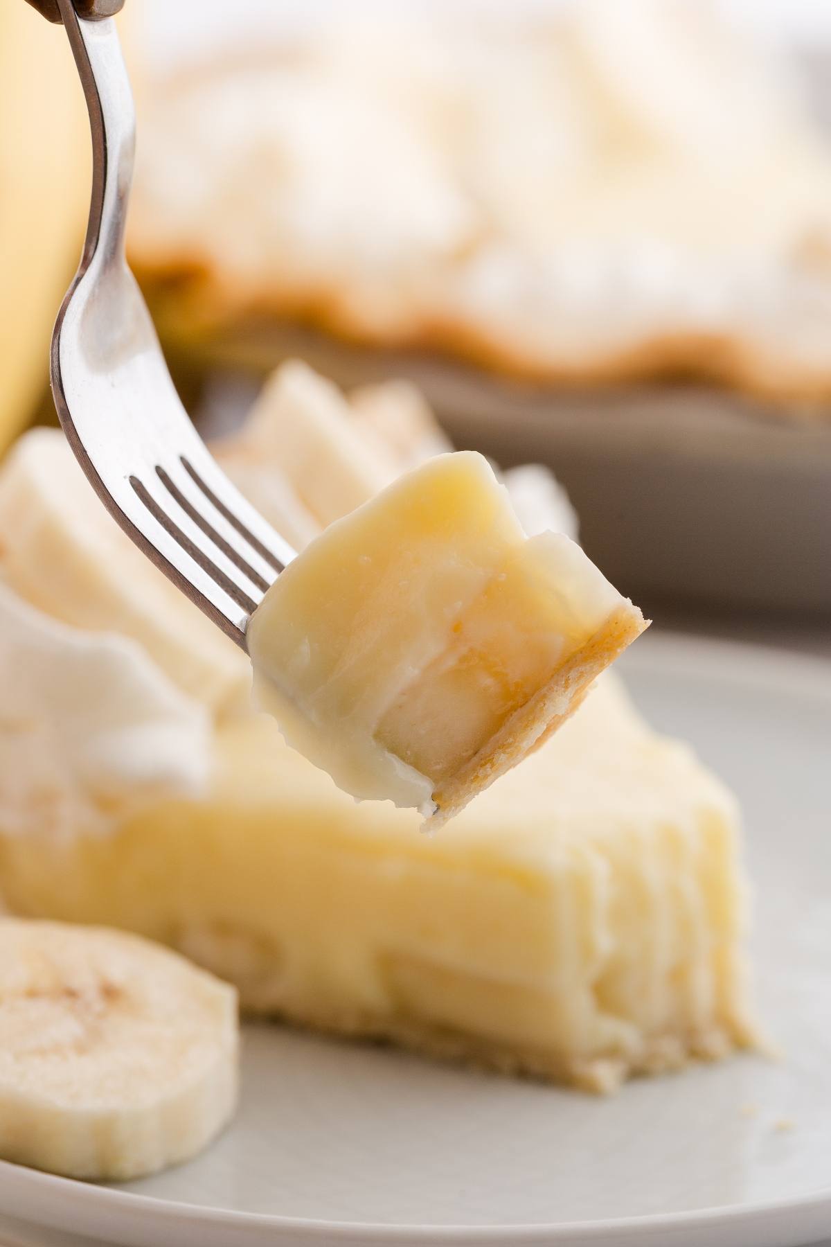 A fork holding a bite of banana cream pie above a slice on a plate, with whipped cream and banana slices.