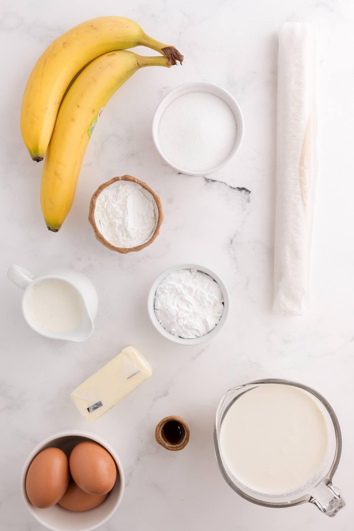 Baking ingredients on a white surface: bananas, eggs, butter, flour, sugar, milk, cream, pastry sheet, and vanilla.