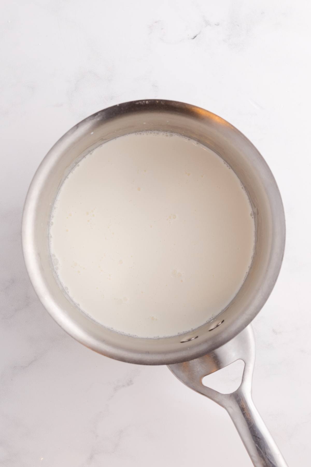 A stainless steel saucepan filled with milk on a white marble surface.