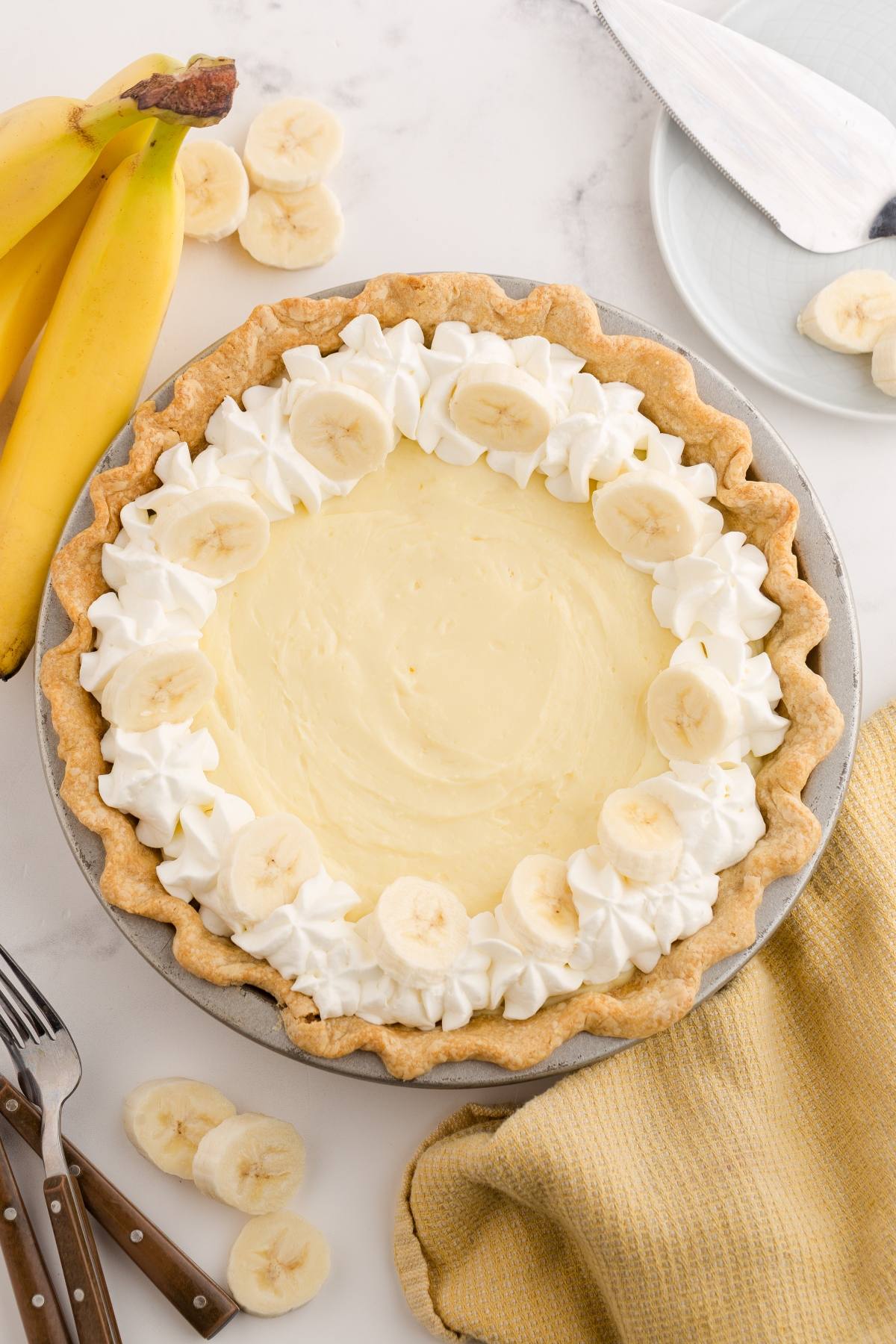 Banana cream pie with whipped cream and banana slices on top, next to whole bananas and a yellow cloth.