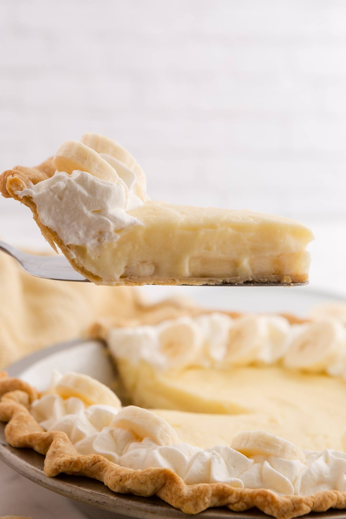 A slice of banana cream pie with whipped cream being lifted from a whole pie in the background.