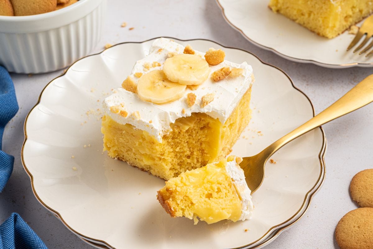 A plate with banana pudding poke cake, whipped topping, banana slices, and a fork with a bite taken out.