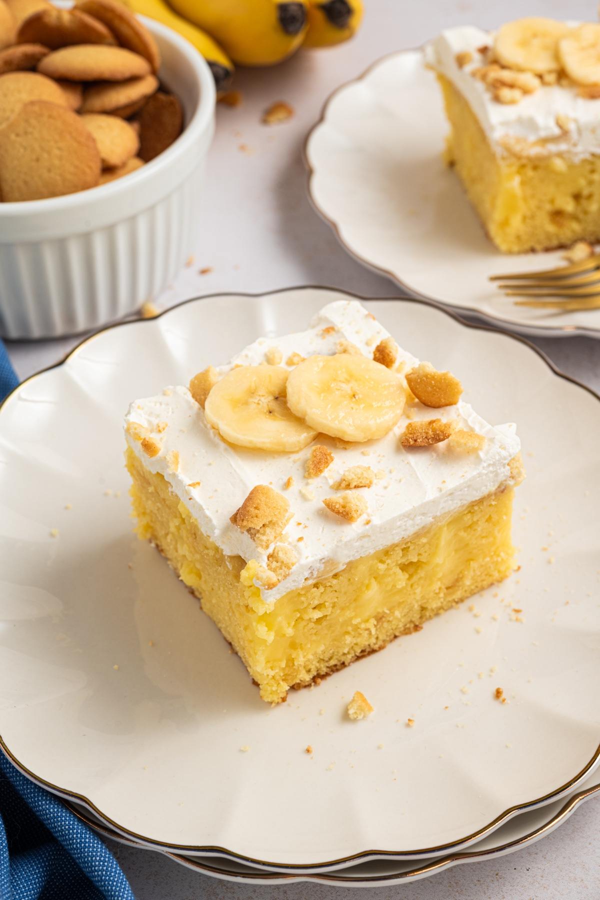 A slice of banana pudding cake topped with whipped cream, banana slices, and cookie crumbs on a white plate.