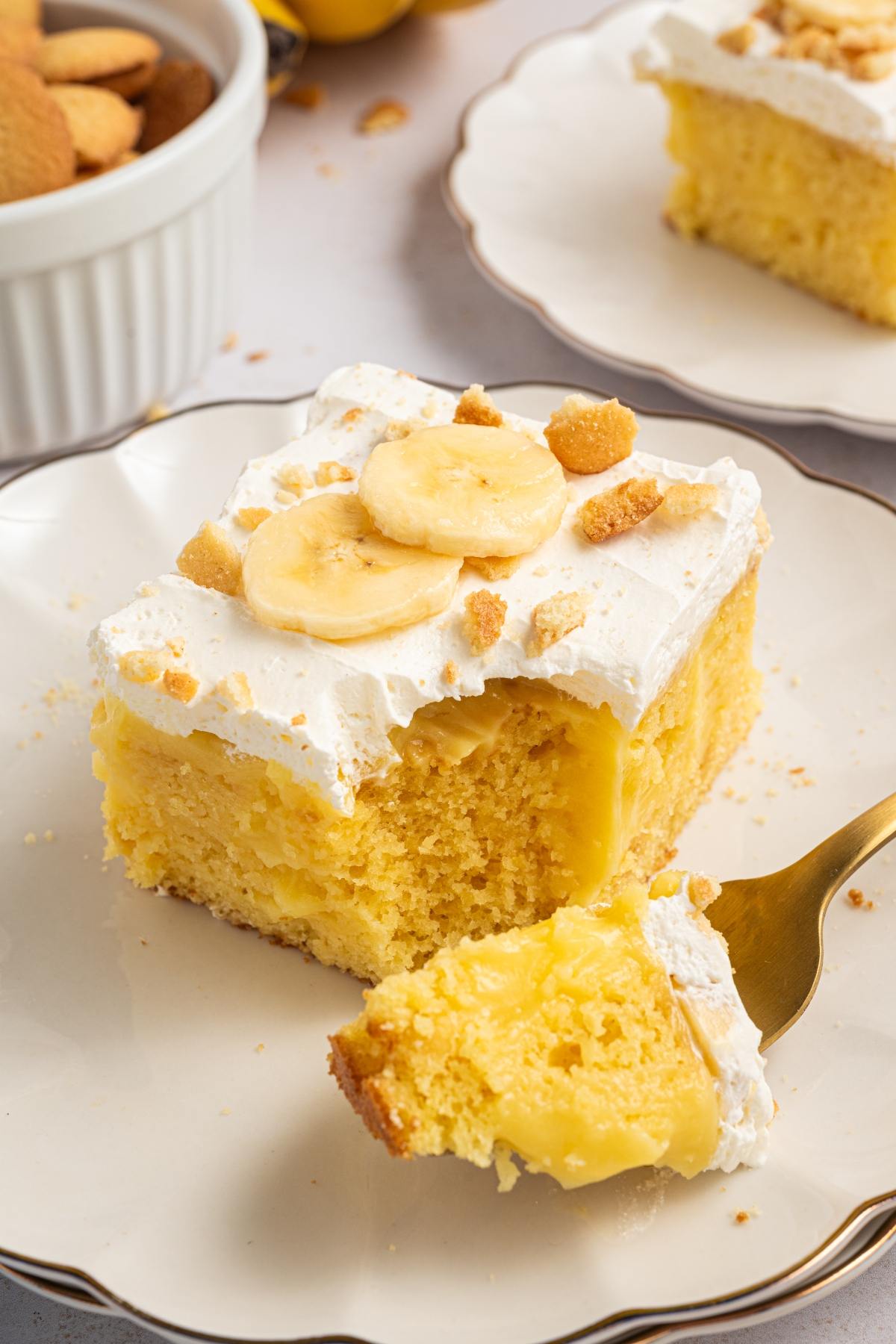 A slice of banana pudding cake with whipped topping, banana slices, and cookie crumbs on a plate with a forkful taken.
