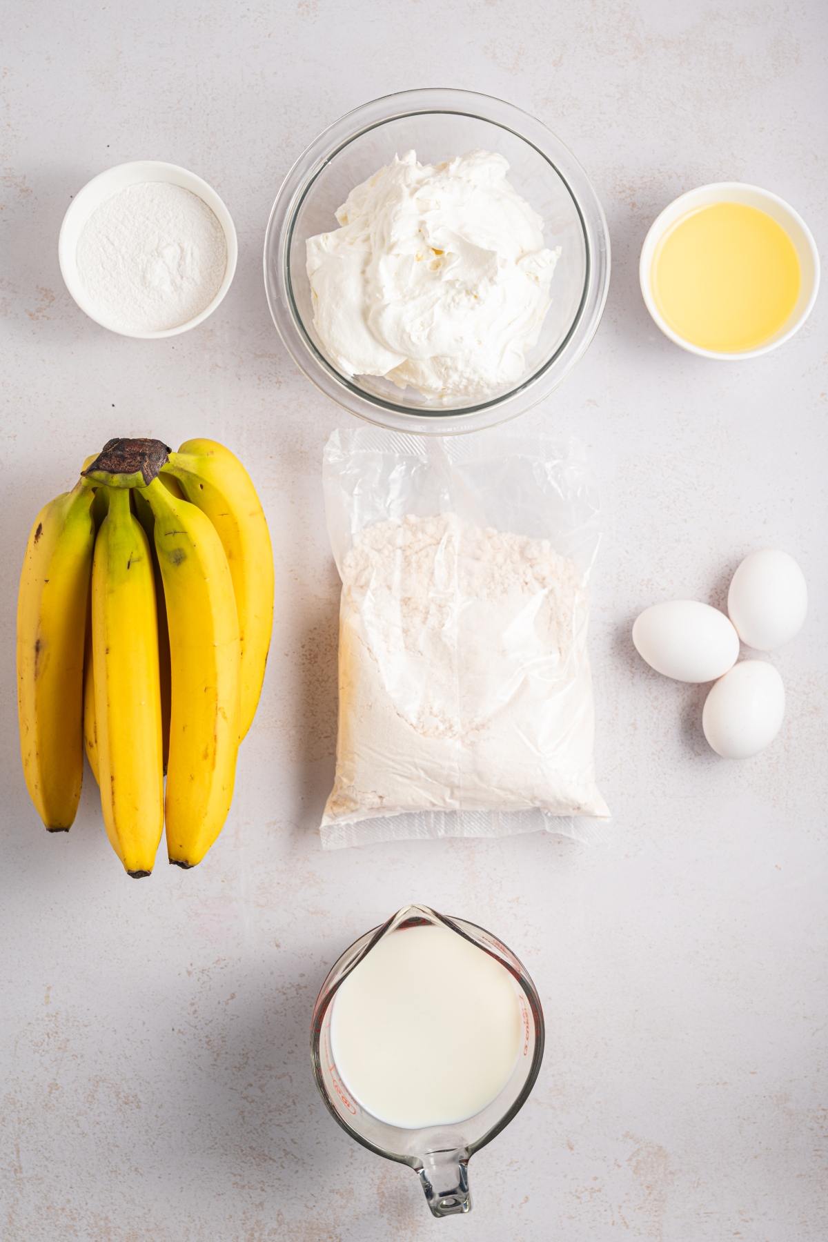 Bananas, eggs, milk, oil, flour, whipped cream, and sugar arranged on a light surface.