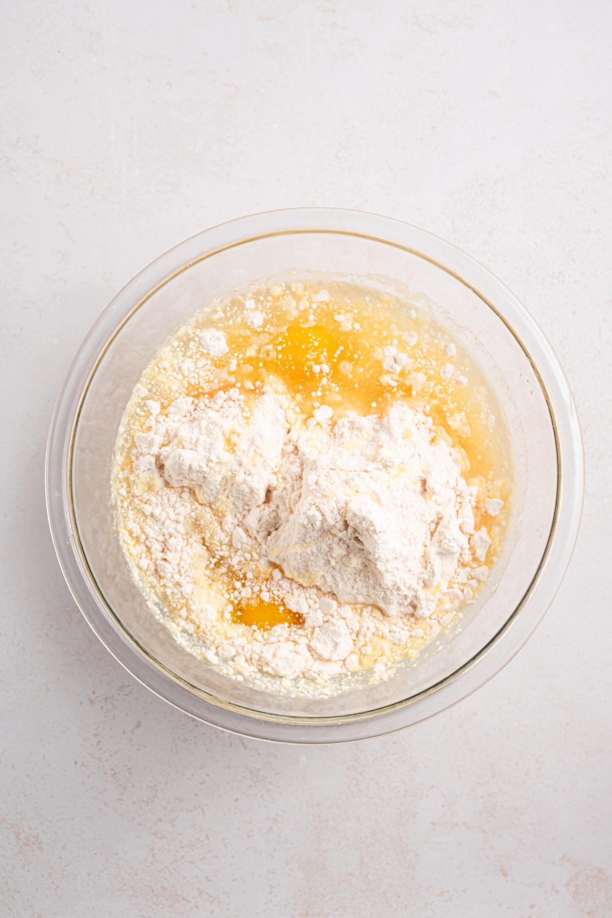 A glass bowl with flour, melted butter, and liquid ingredients on a light surface, ready to be mixed.