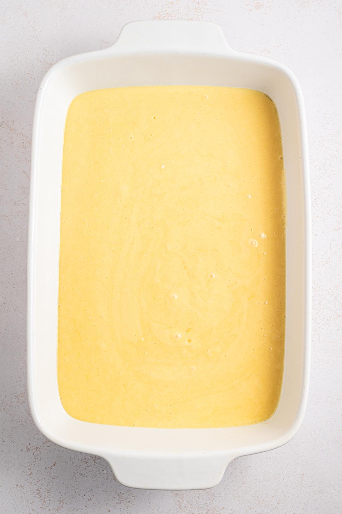 White rectangular baking dish filled with smooth yellow cornbread batter on a light surface.