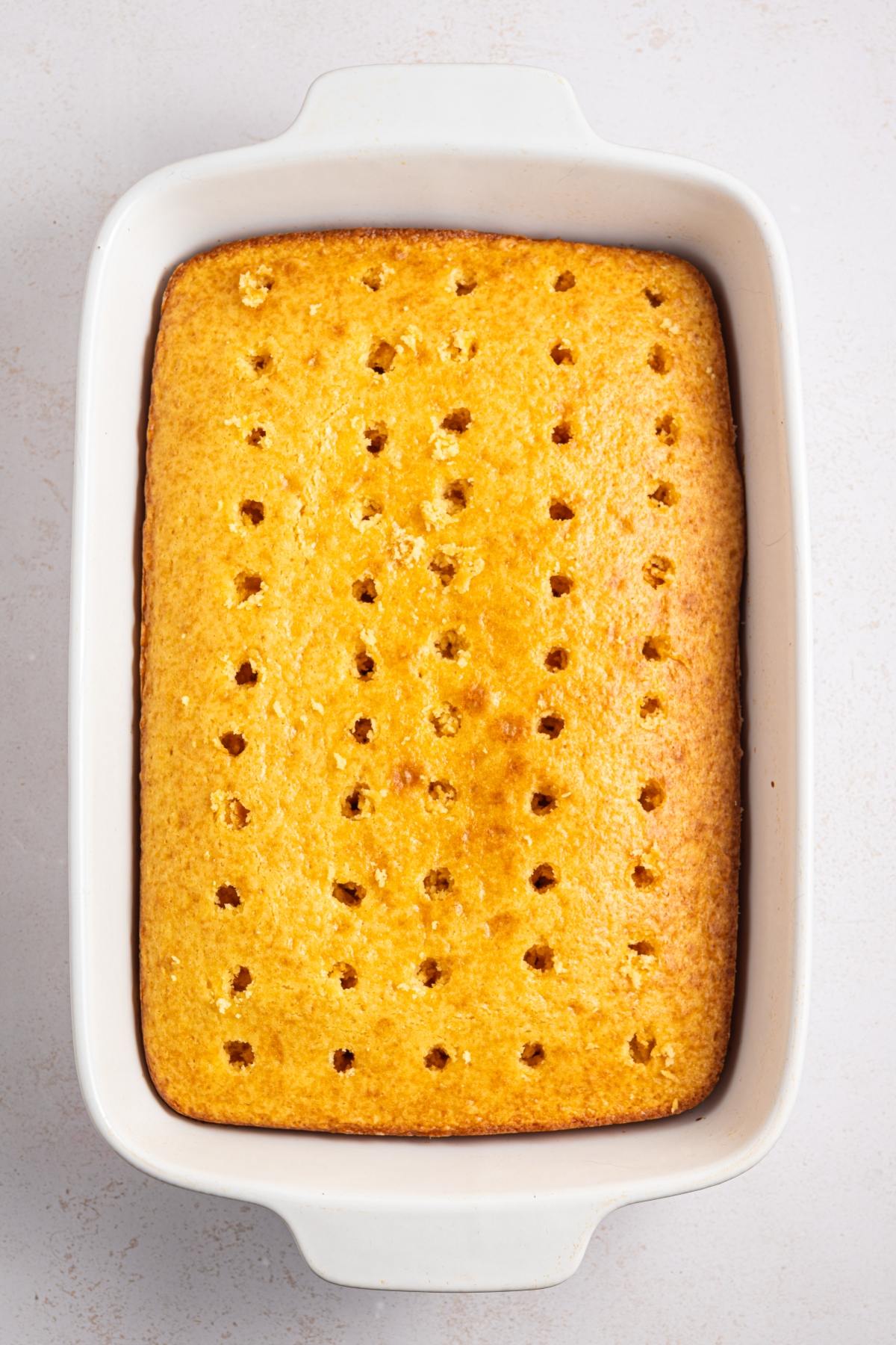 Golden sheet cake with evenly spaced holes poked on top, in a white rectangular baking dish.