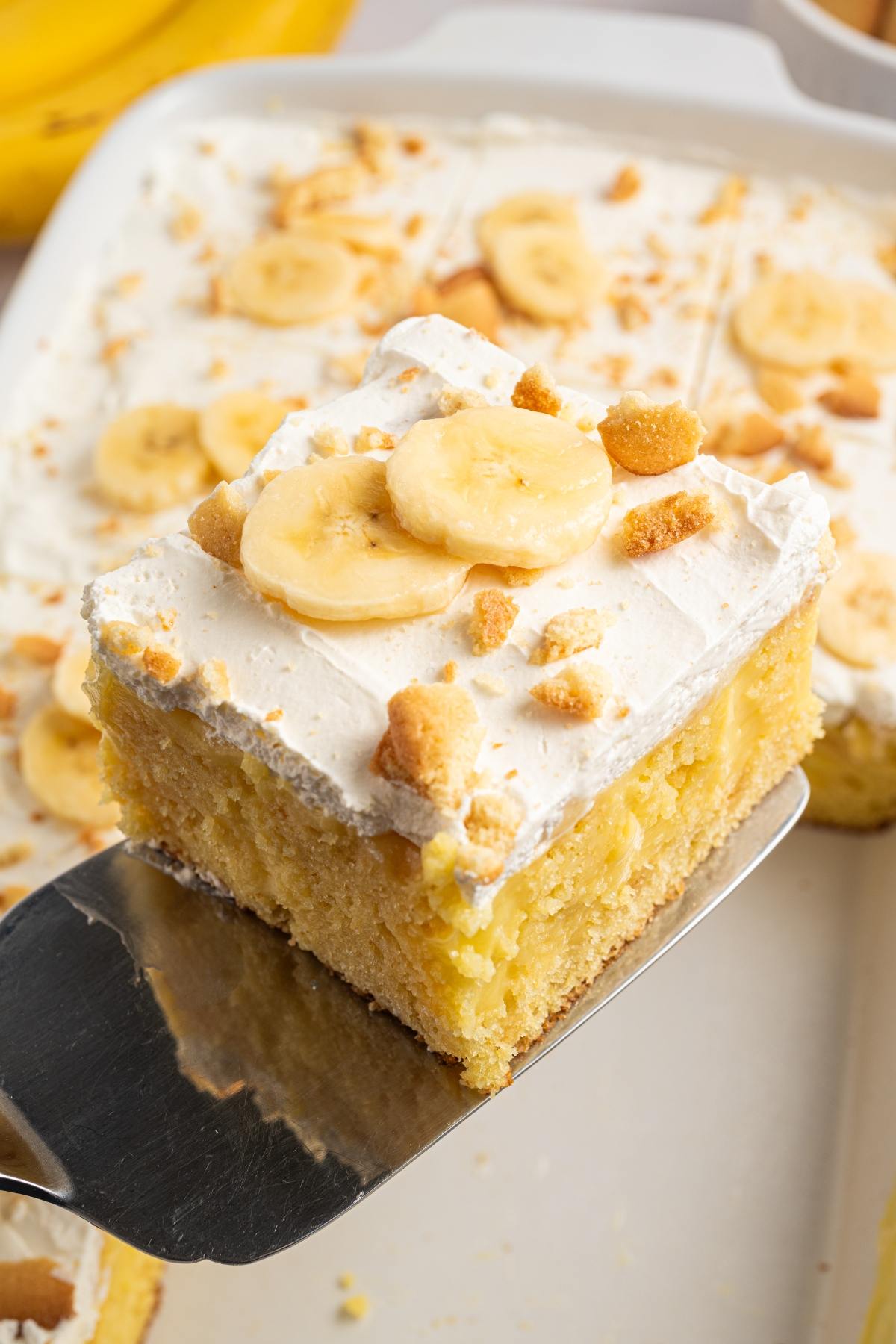 A slice of banana pudding cake with whipped topping, banana slices, and cookie crumbles on a spatula.