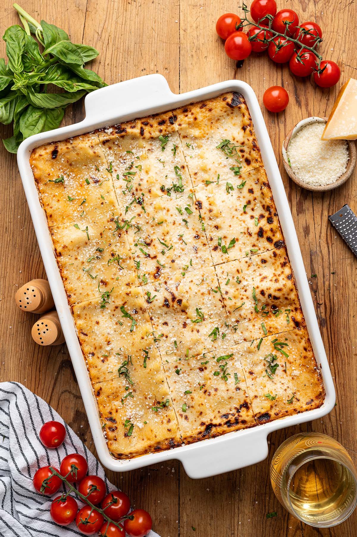 Baked lasagna alla Bolognese in a white dish, topped with herbs, surrounded by tomatoes, basil, cheese, and a glass of wine.