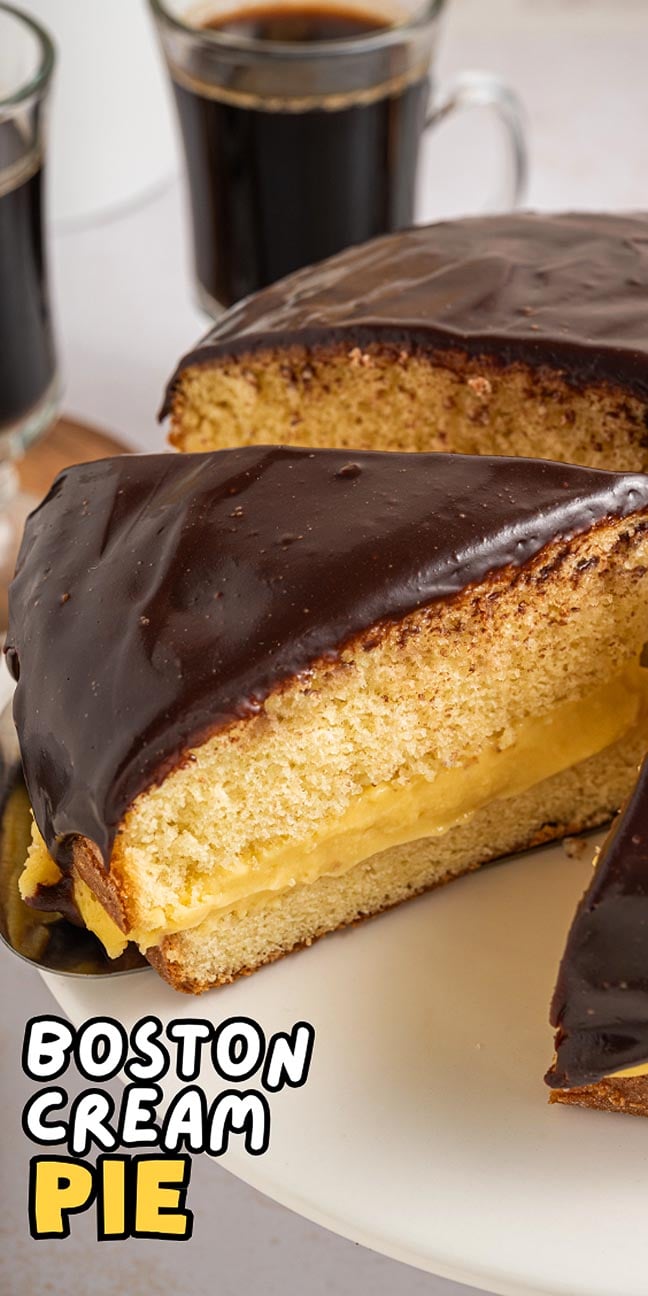 A sliced Boston cream pie with rich chocolate glaze and creamy yellow custard filling is served on a plate alongside a cup of coffee.