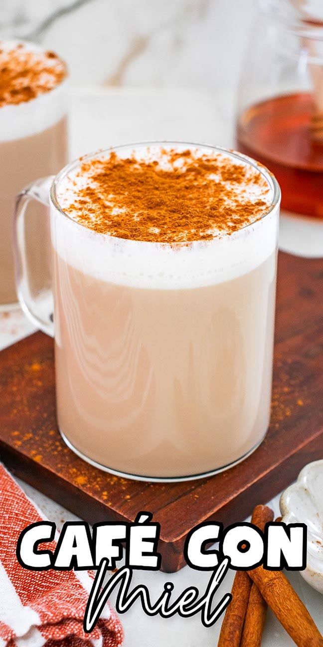 A glass mug of café con miel topped with cinnamon sits on a wooden board, surrounded by honey and fragrant cinnamon sticks.