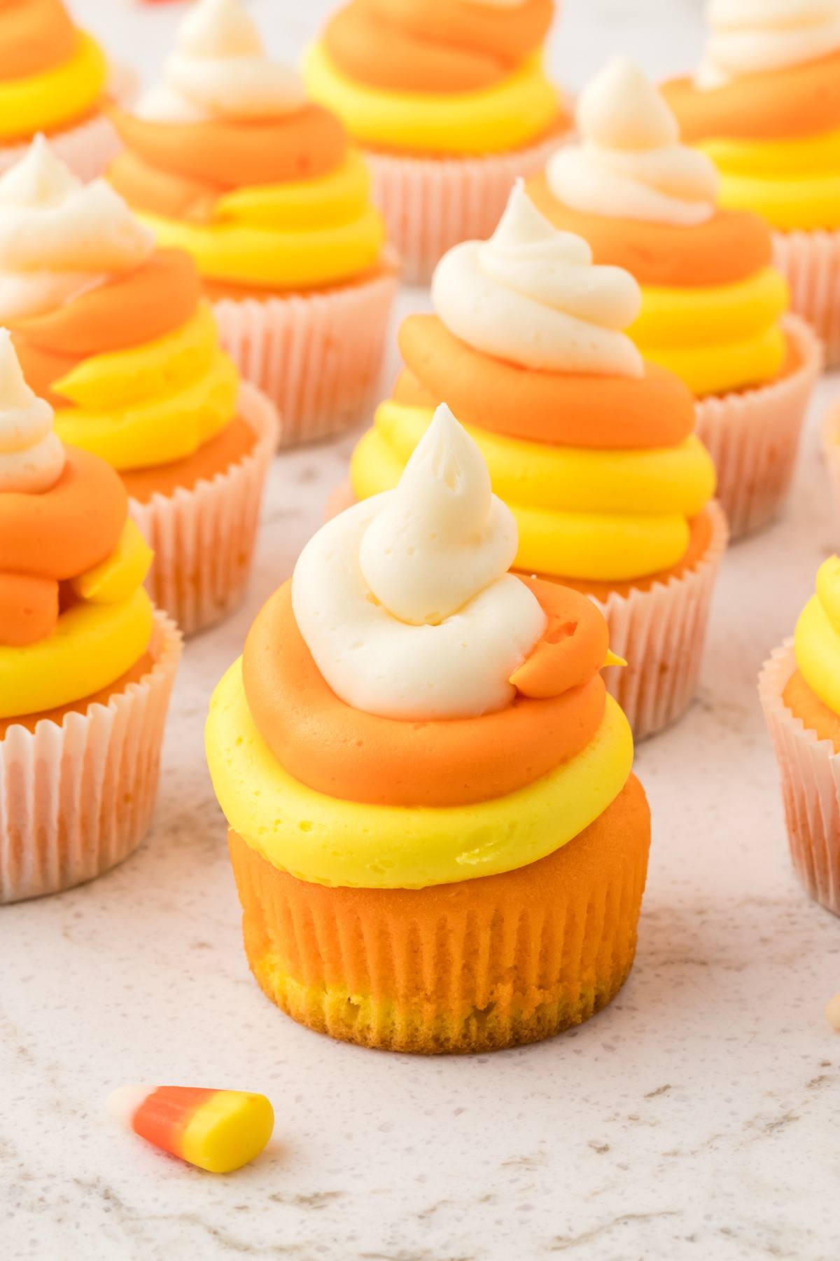 Cupcakes with yellow, orange, and white frosting swirled to resemble candy corn, with a candy corn piece in front.