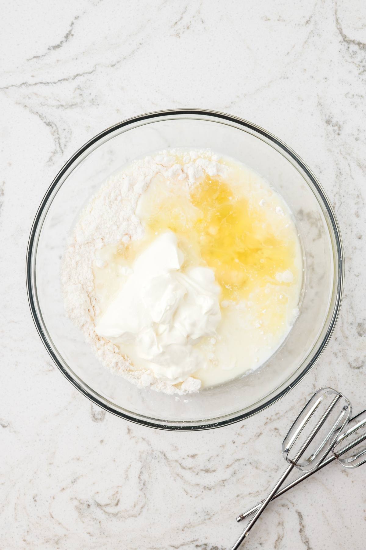 A glass bowl with flour, yogurt, eggs, and milk, next to metal beaters on a marble countertop.