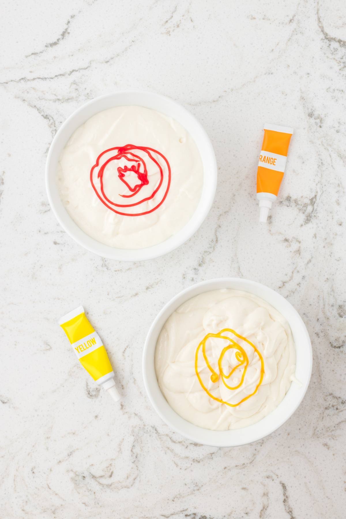 Two bowls of white frosting with red and yellow food coloring, and tubes labeled orange and yellow beside them.