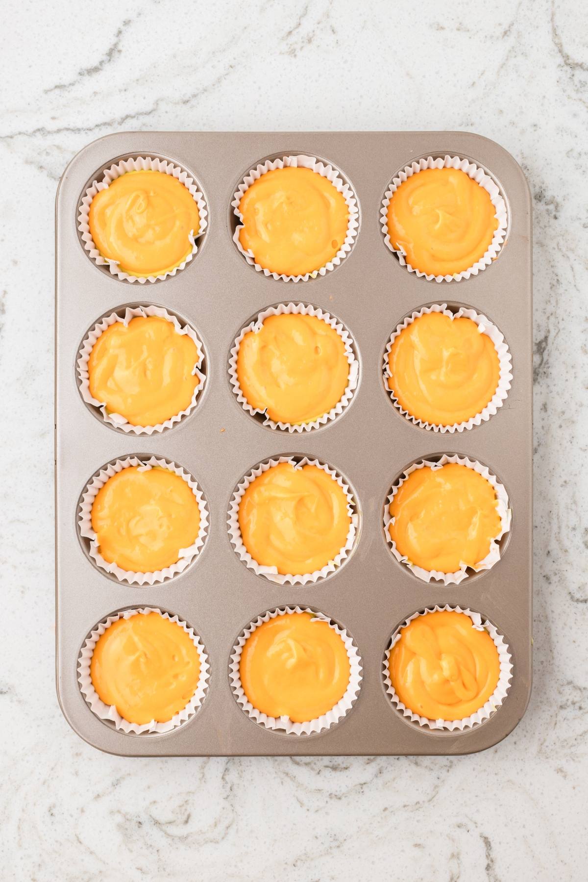 Muffin tin with 12 cupcake liners filled with orange batter on a light marble surface.