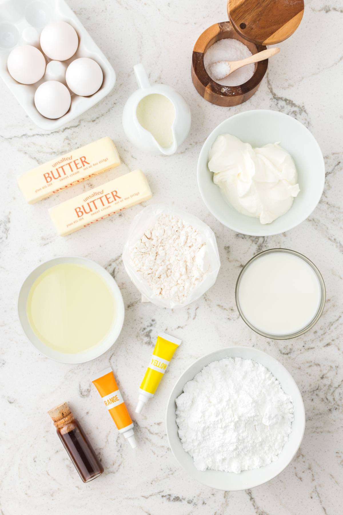 Baking ingredients on a counter: eggs, butter, flour, sugar, milk, vanilla, oil, salt, food coloring, and cream.