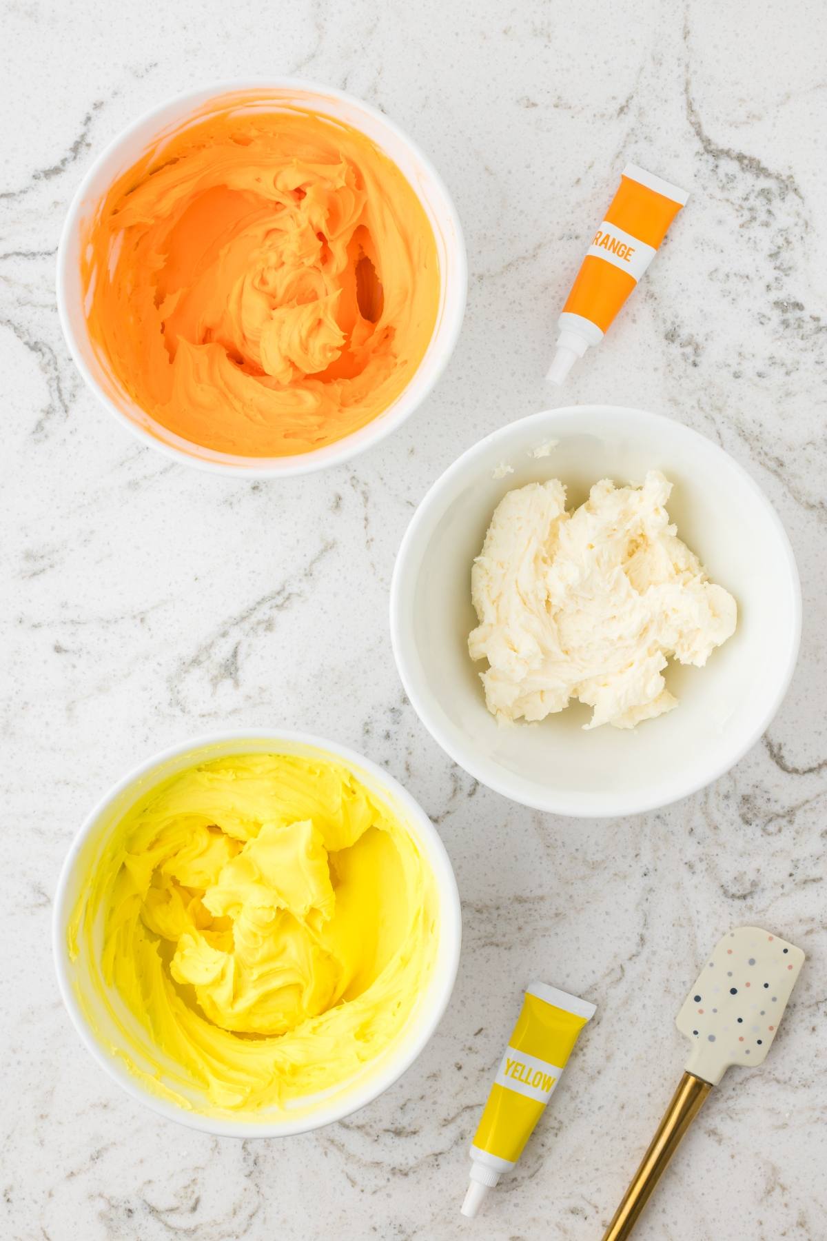 Three bowls of frosting (orange, white, yellow) with matching food coloring tubes on a white marble surface.
