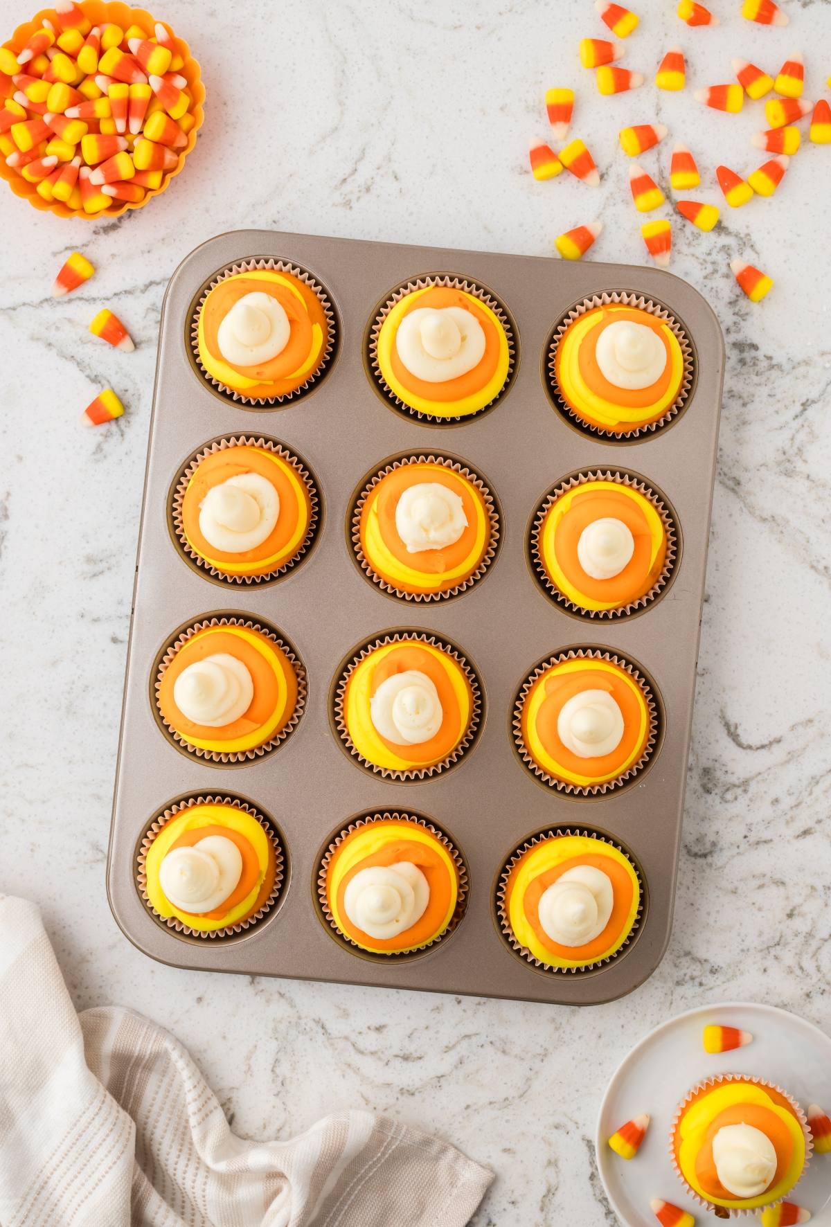 A muffin tin with 12 orange and yellow cupcakes, topped with white frosting, on a marble surface.