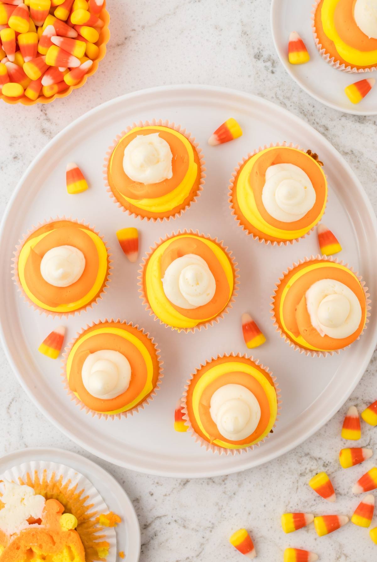 Eight candy corn-inspired cupcakes with orange and yellow frosting on a white plate, surrounded by candy corn.
