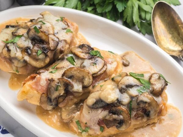 Three pieces of chicken Valdostana topped with mushrooms and melted cheese on a white plate with creamy sauce.
